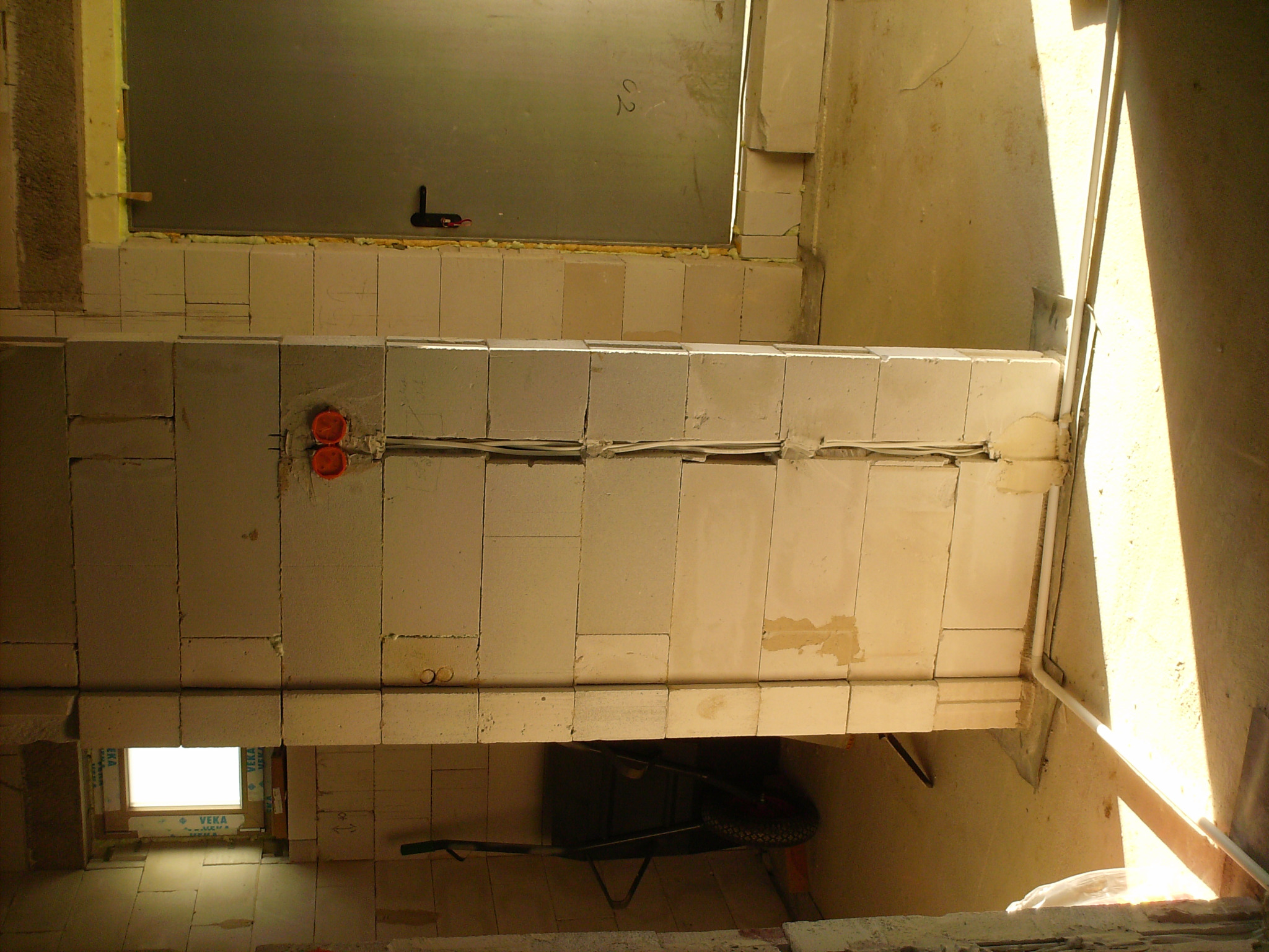 Wewnętrzna ściana z pustaków z widocznymi puszkami elektrycznymi i przewodami w trakcie montażu, z wózkiem budowlanym w tle i oknem z lewej strony.