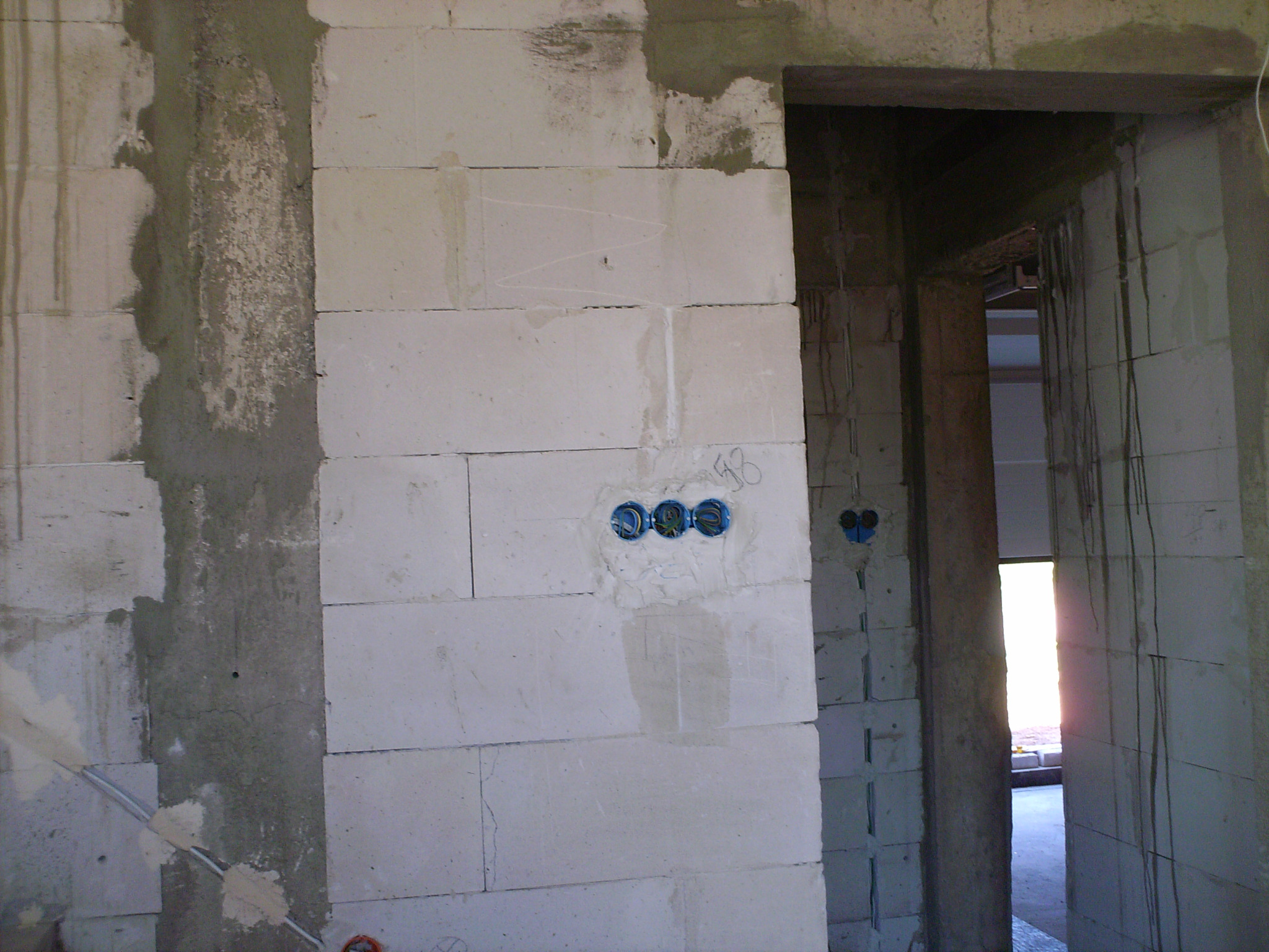 Surowa ściana z betonu komórkowego w trakcie prac instalacyjnych, widoczne puszki elektryczne z przewodami, ślady zaprawy i instalacji elektrycznej w nowo budowanym pomieszczeniu.