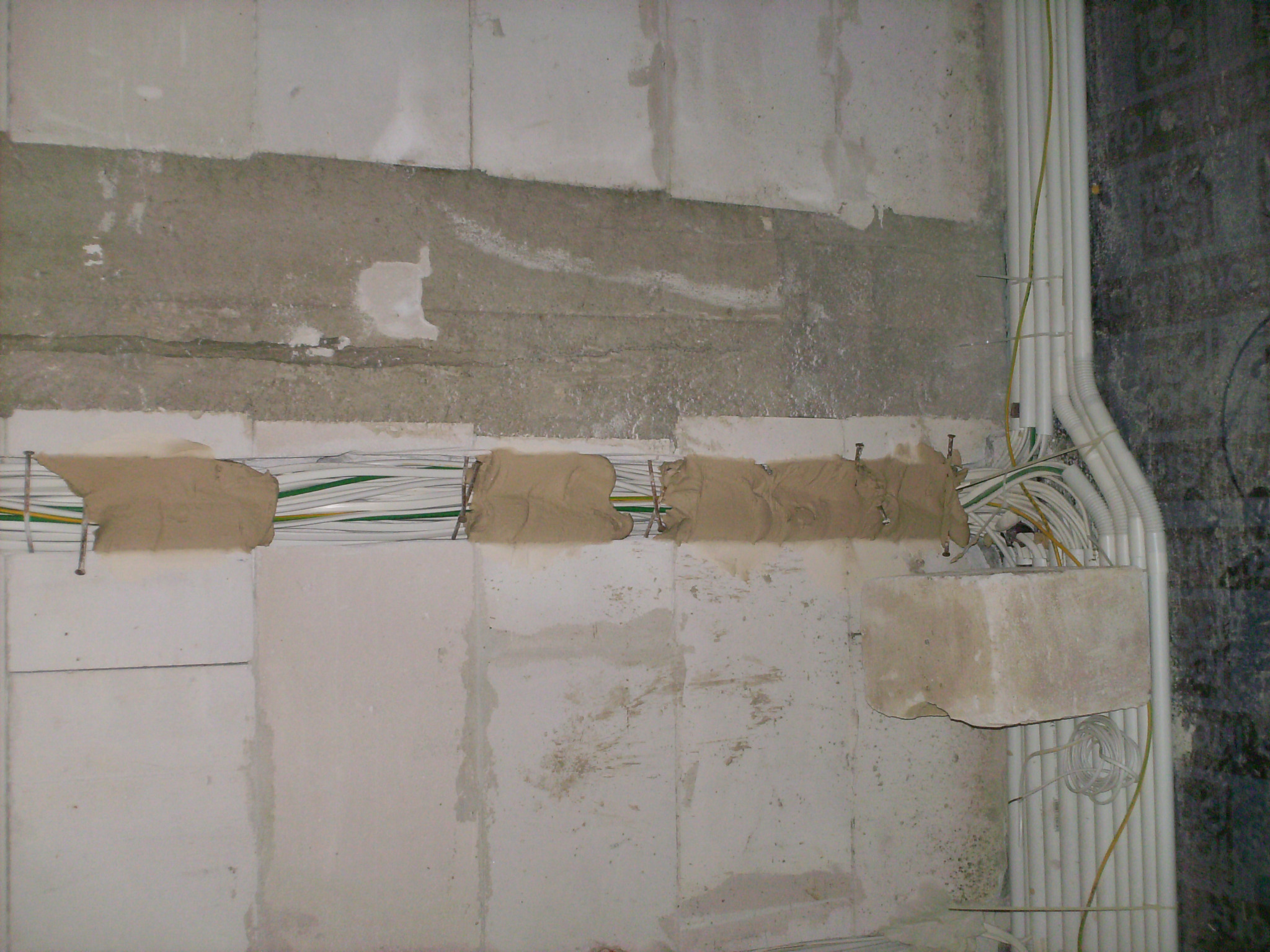 Surowe ściany z betonu komórkowego z częściowo zamaskowanymi przewodami elektrycznymi, zabezpieczonymi gipsem, oraz wiązką rur osłonowych pionowo ułożonych po prawej stronie.