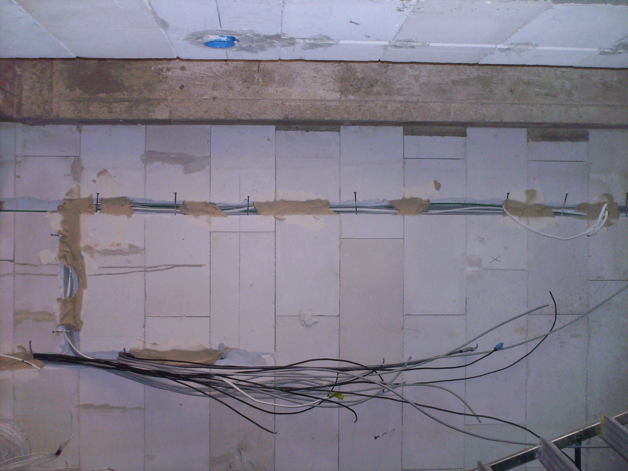 Widok z góry na surowy strop z instalacją elektryczną w trakcie montażu, kable przymocowane do bloczków z betonu komórkowego za pomocą gwoździ, widoczna puszka instalacyjna w suficie.
