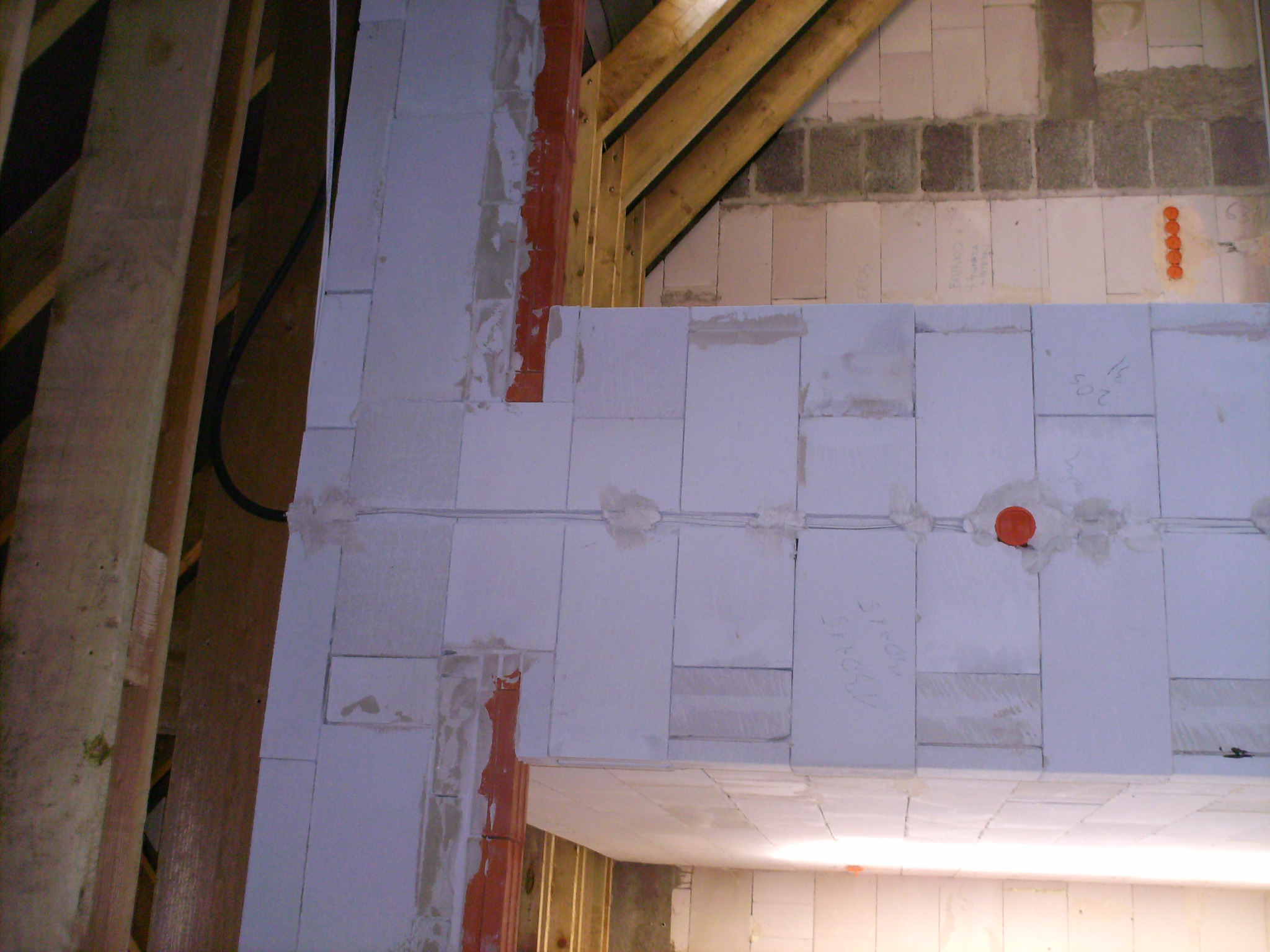 Wewnętrzna ściana z białych bloczków w trakcie budowy, widoczne kable elektryczne w bruzdach, elementy konstrukcji drewnianej dachu, fragment instalacji elektrycznej w ścianie.