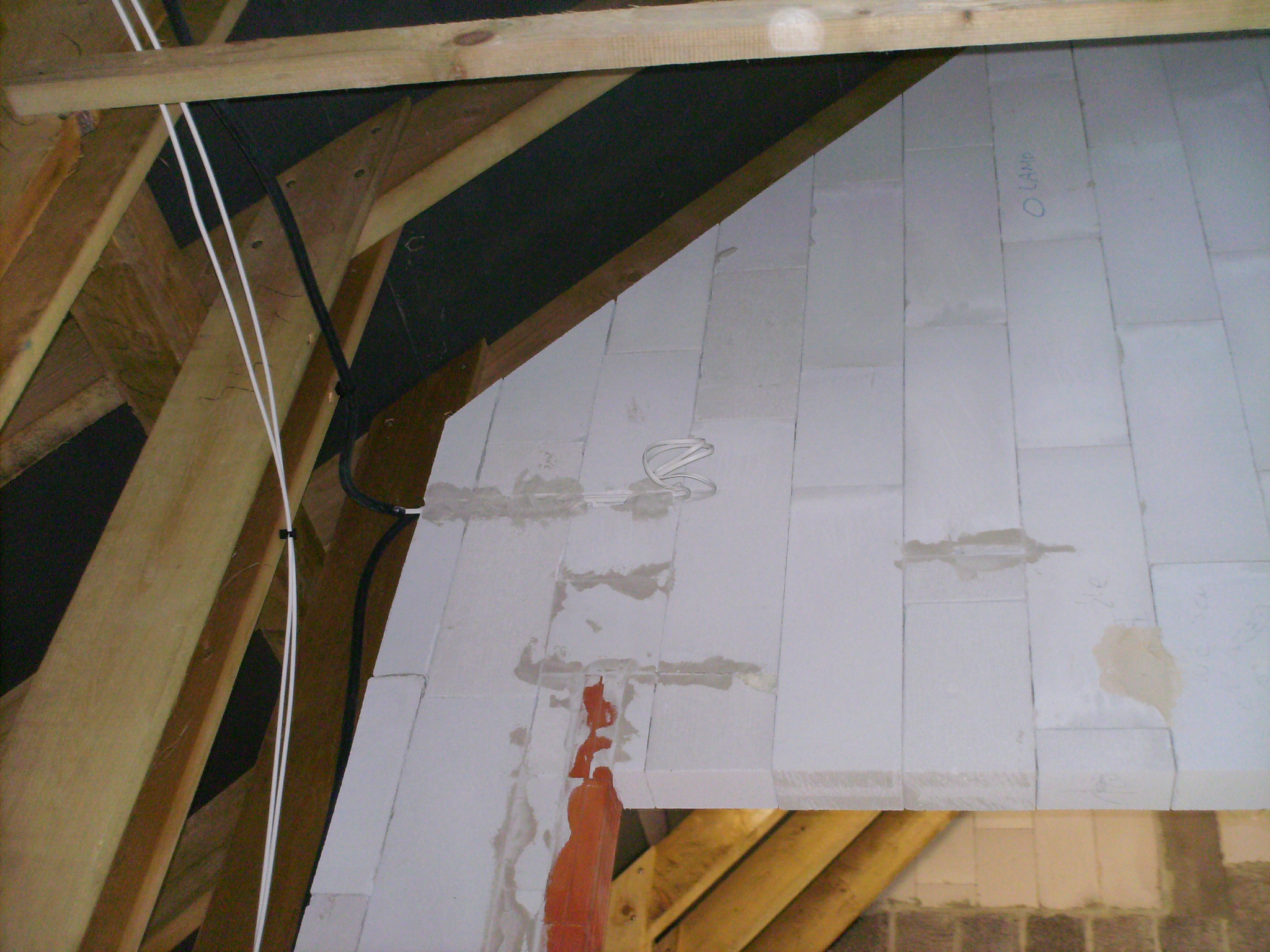 Poddasze w trakcie izolacji, widoczne kable elektryczne przymocowane do drewnianej konstrukcji dachu oraz białe panele izolacyjne z widocznymi śladami kleju i oznaczeniem 'O LAMP'.