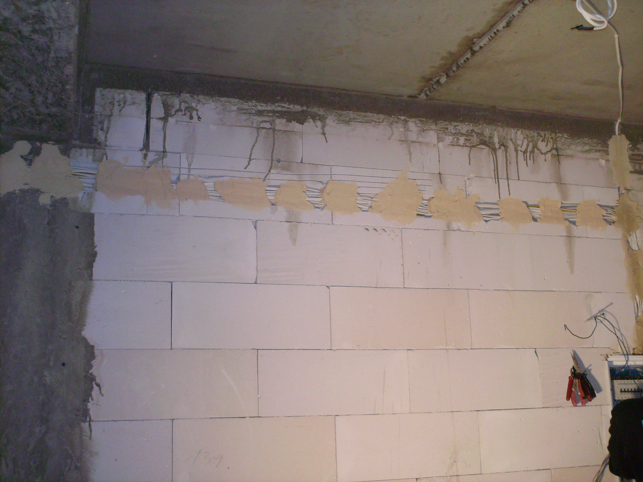 Ściana z betonu komórkowego z wyciętymi bruzdami pod przewody elektryczne, częściowo zamaskowanymi gipsem, widoczne narzędzia i skrzynka elektryczna.