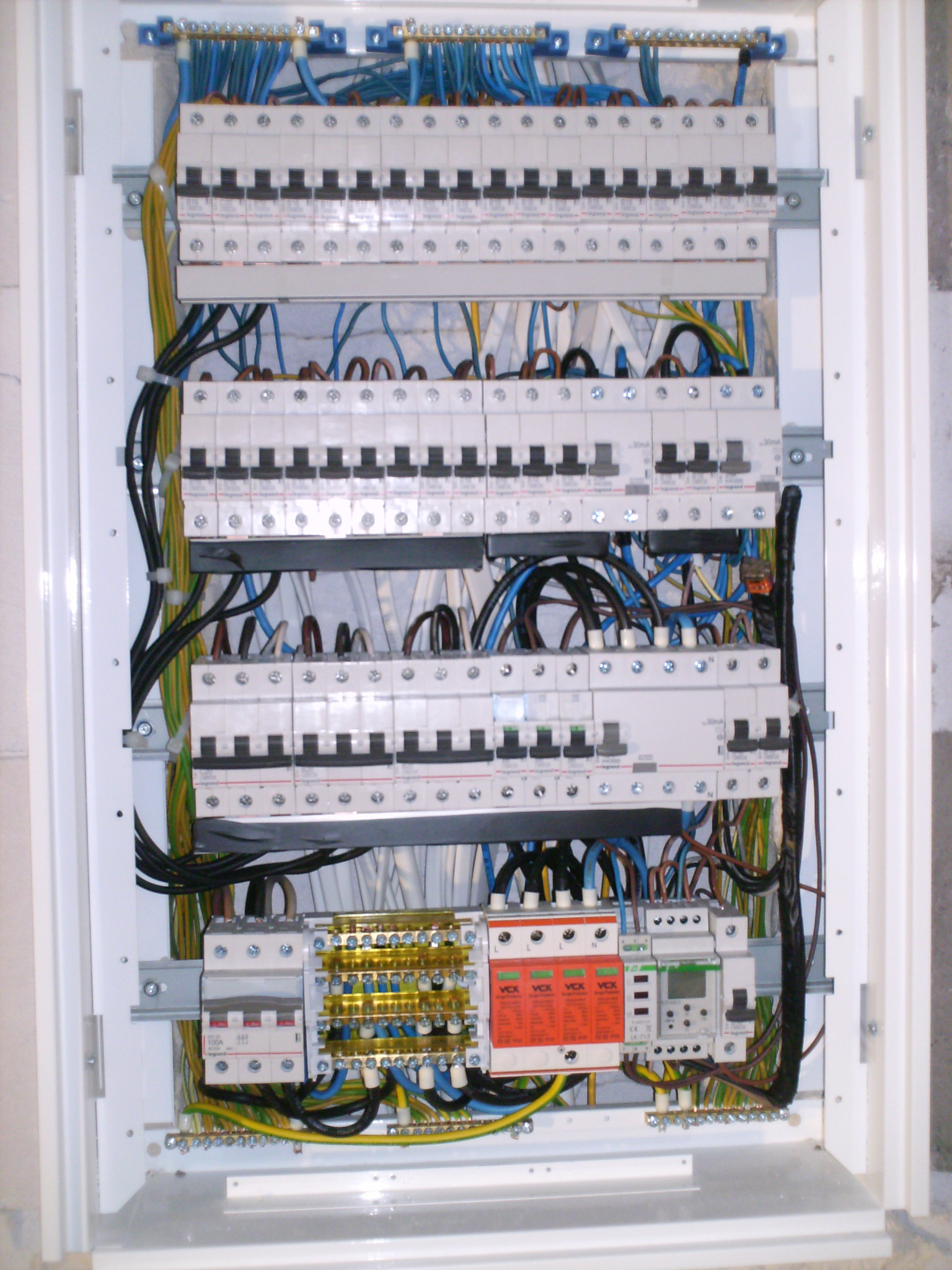 Rozdzielnica elektryczna w obudowie, widoczne zabezpieczenia różnicowoprądowe, bezpieczniki, listwy zaciskowe, przewody w różnych kolorach, ułożone wewnątrz białej obudowy, zamontowanej na ścianie.