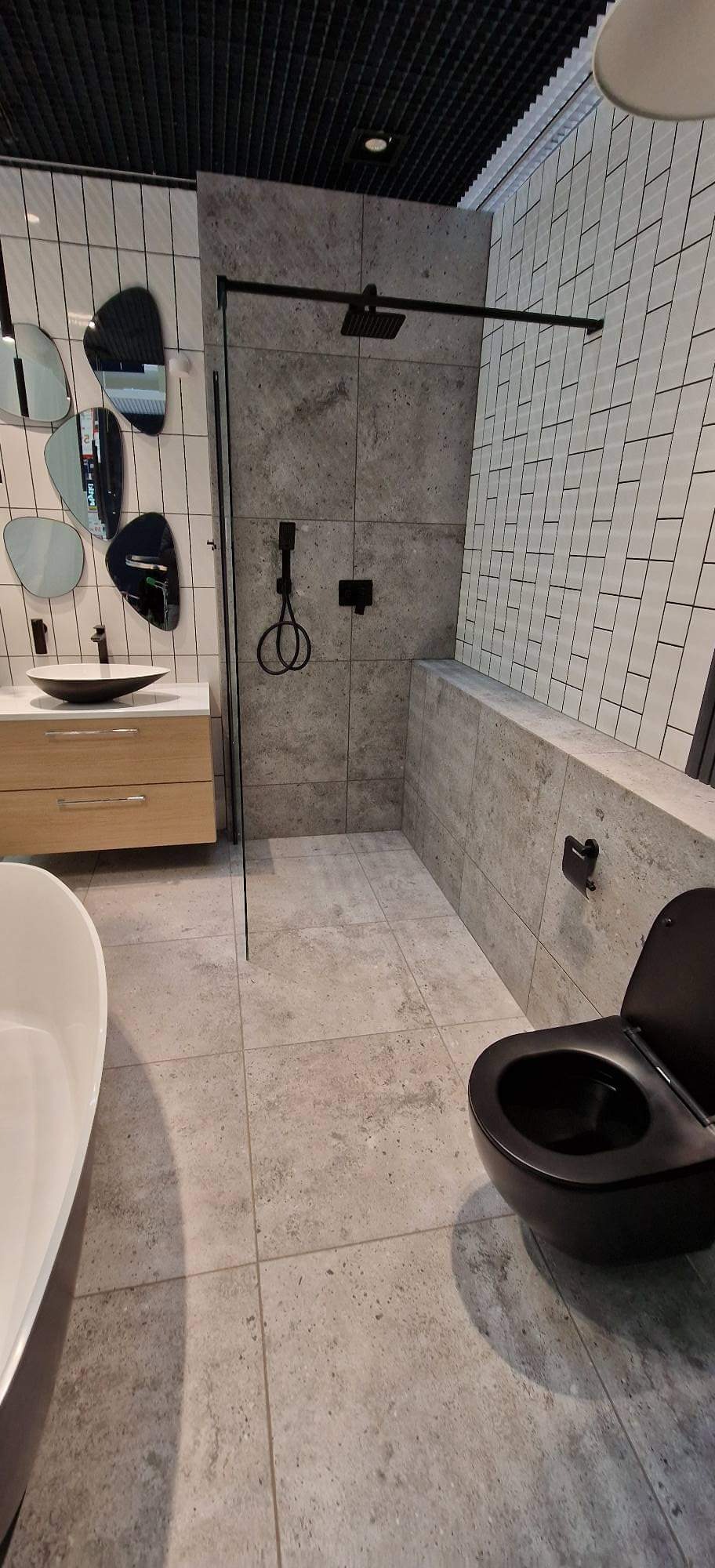 Nowoczesna łazienka w szarościach z czarnymi akcentami: kabina prysznicowa typu walk-in, umywalka nablatowa z szafką z jasnego drewna, czarna toaleta i asymetryczne lustra na ścianie.