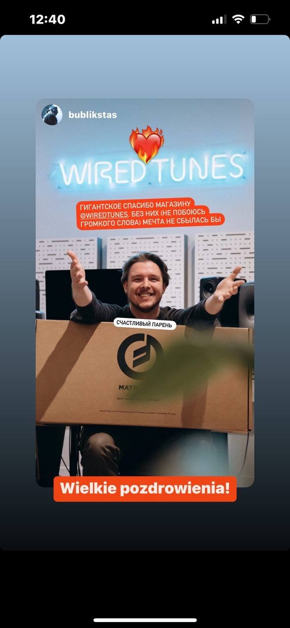 Uśmiechnięty mężczyzna trzyma pudełko z logo firmy MATĘ na tle neonowego napisu WIRED TUNES i głośników studyjnych.