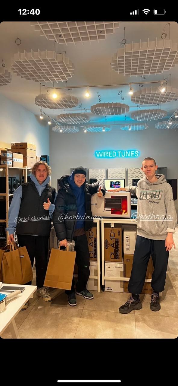 Trzech młodych mężczyzn pozuje z torbami zakupowymi w sklepie z nagłośnieniem Wired Tunes, przed regałem z głośnikami CLA-10 Active i neonowym logo firmy.