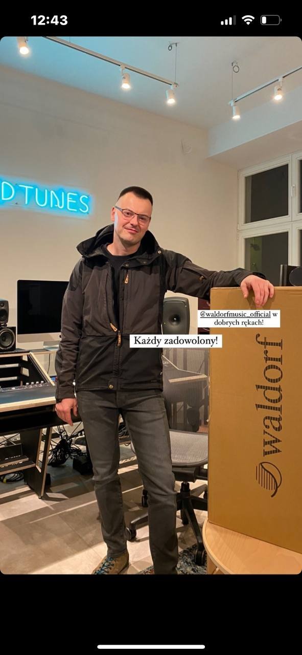 Mężczyzna w okularach stoi w studiu nagraniowym obok sprzętu Waldorf, w tle neonowy napis 'D TUNES'.