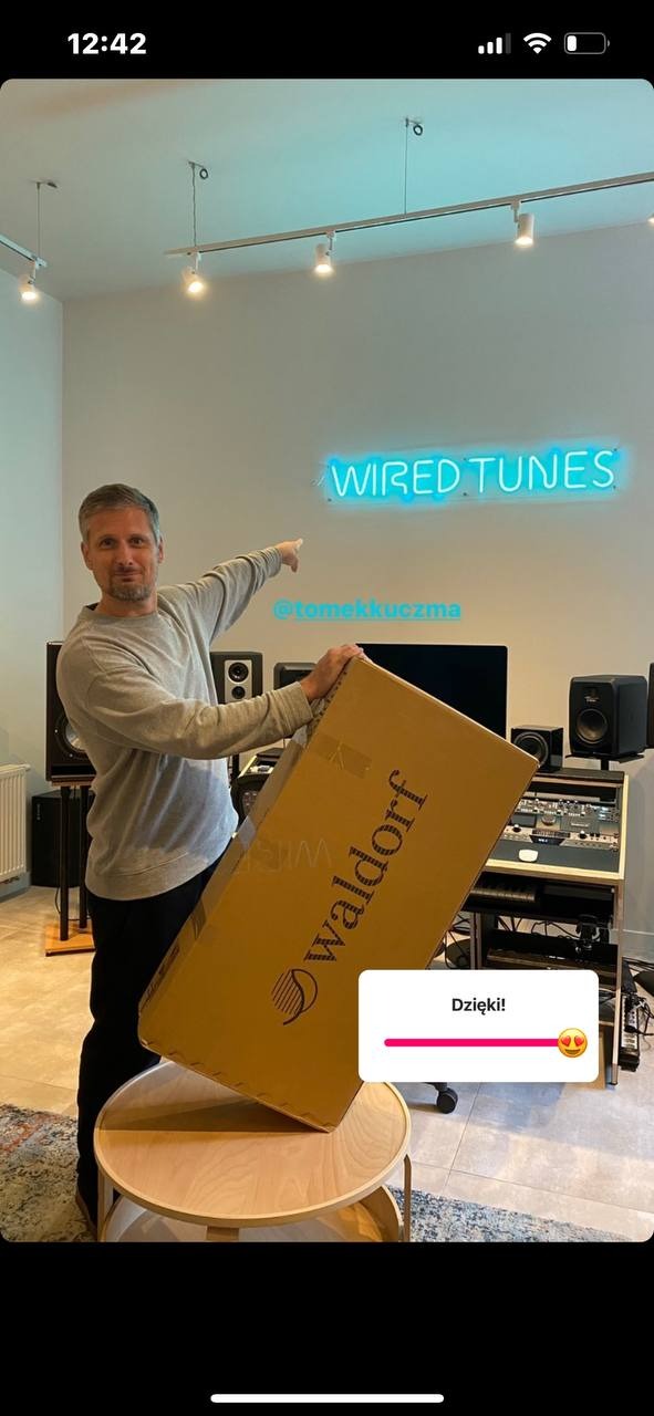 Mężczyzna wskazujący na neonowy napis 'Wired Tunes' i trzymający karton z napisem 'Waldorf' w studiu nagraniowym z widocznym sprzętem audio.