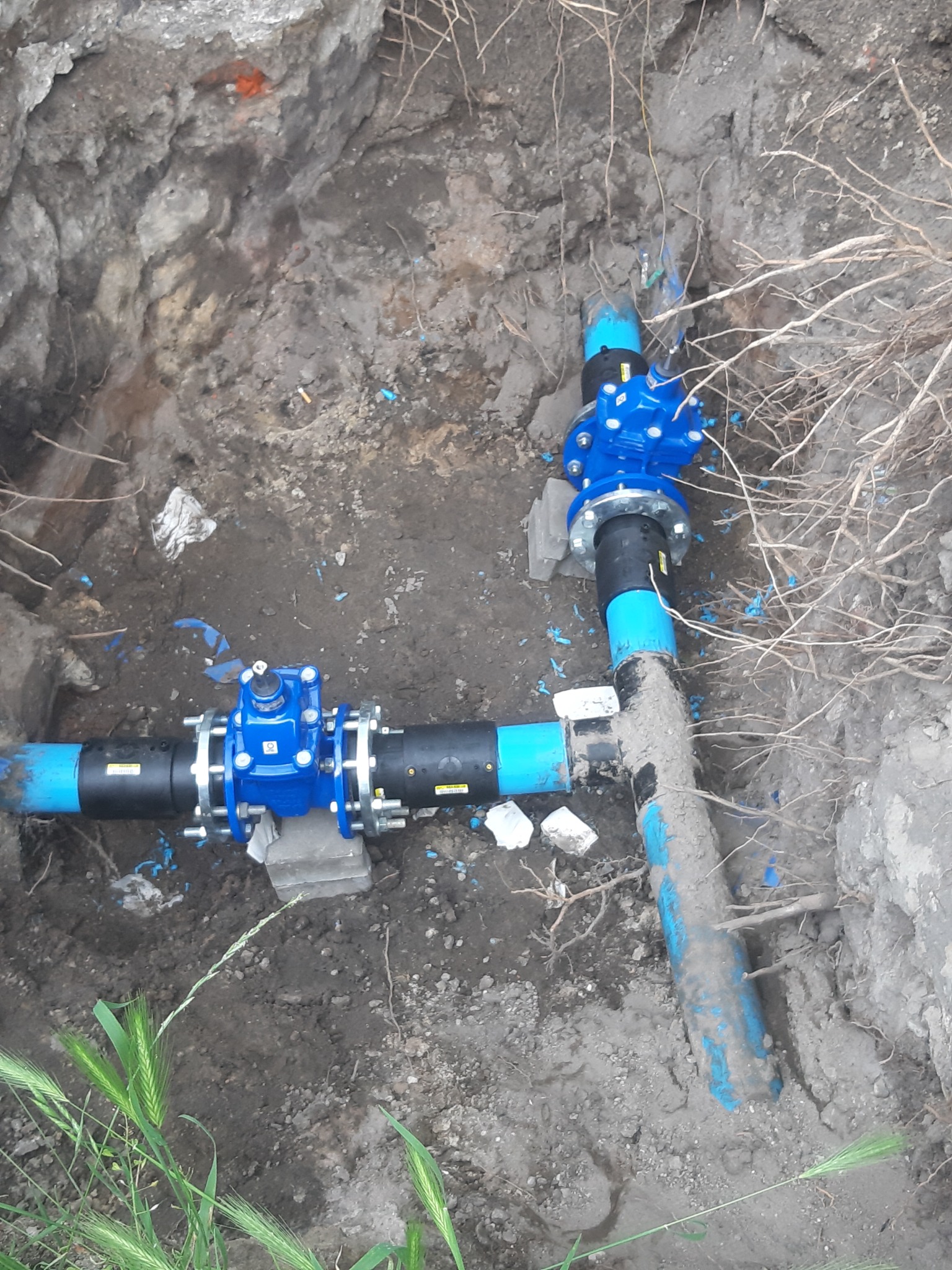 Niebieskie zawory na czarnych rurach w wykopie ziemnym, prawdopodobnie podczas prac związanych z instalacją wodociągową w Bieruniu. Widoczne ślady zabrudzeń ziemią.