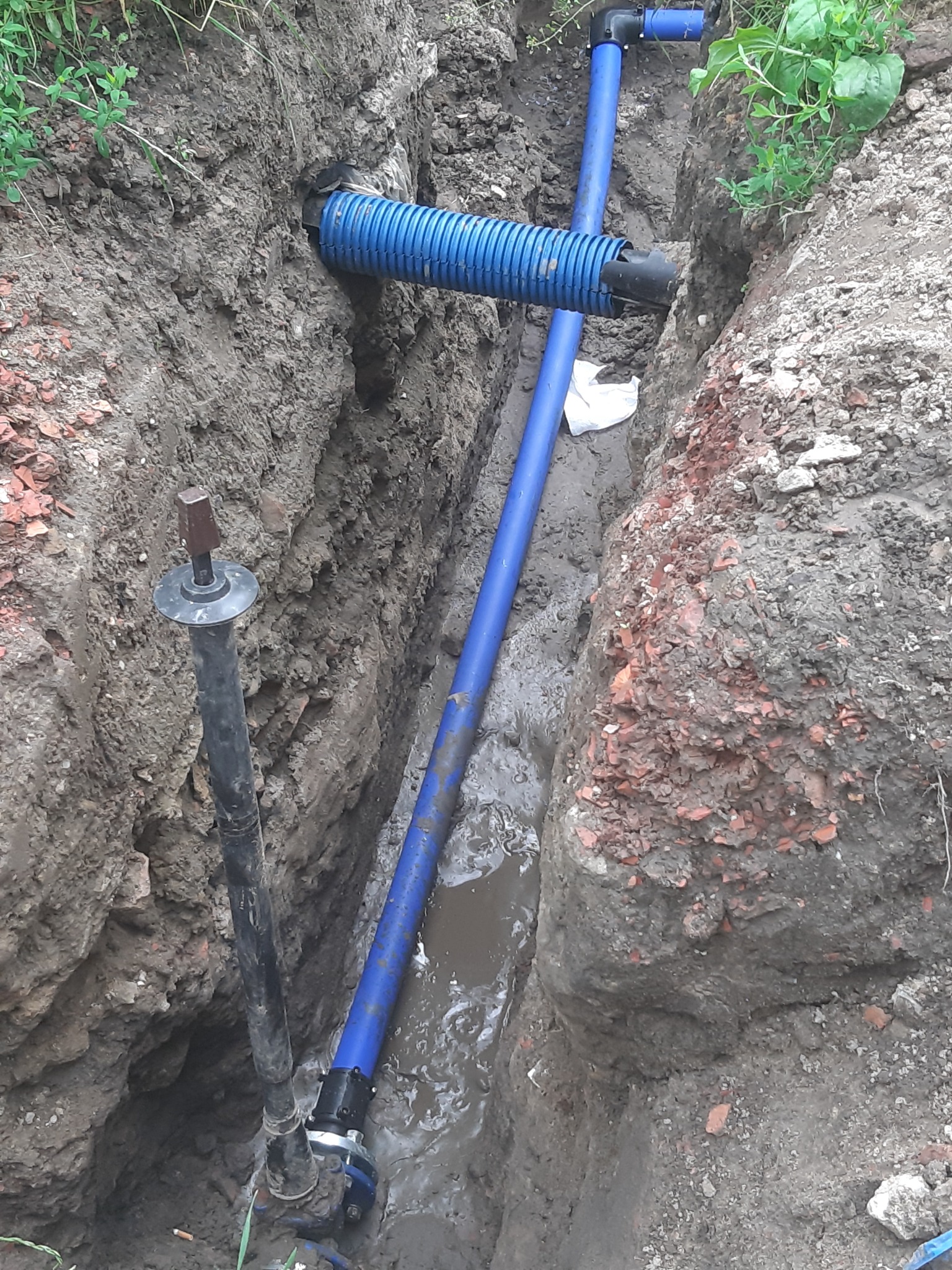 Wykop z ułożonymi niebieskimi rurami wodociągowymi i czarną rurą pionową z metalową nasadką, częściowo wypełniony wodą, widoczne ślady ziemi i gruzu.