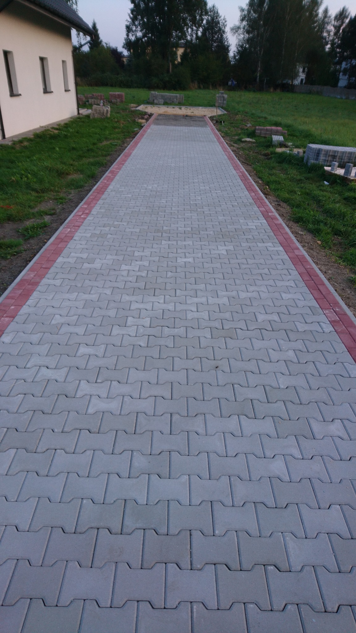 Ułożony chodnik z szarej kostki brukowej w kształcie litery H, obramowany czerwoną kostką, prowadzący do domu z jasną elewacją i zielonym trawnikiem w tle.