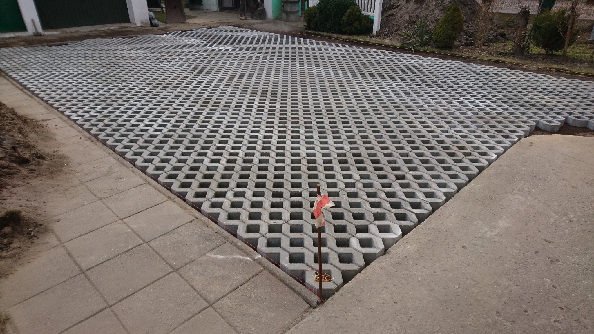 Ułożona szara ażurowa nawierzchnia na podjeździe, widoczny słupek geodezyjny z czerwoną taśmą, obrzeża z kostki brukowej.