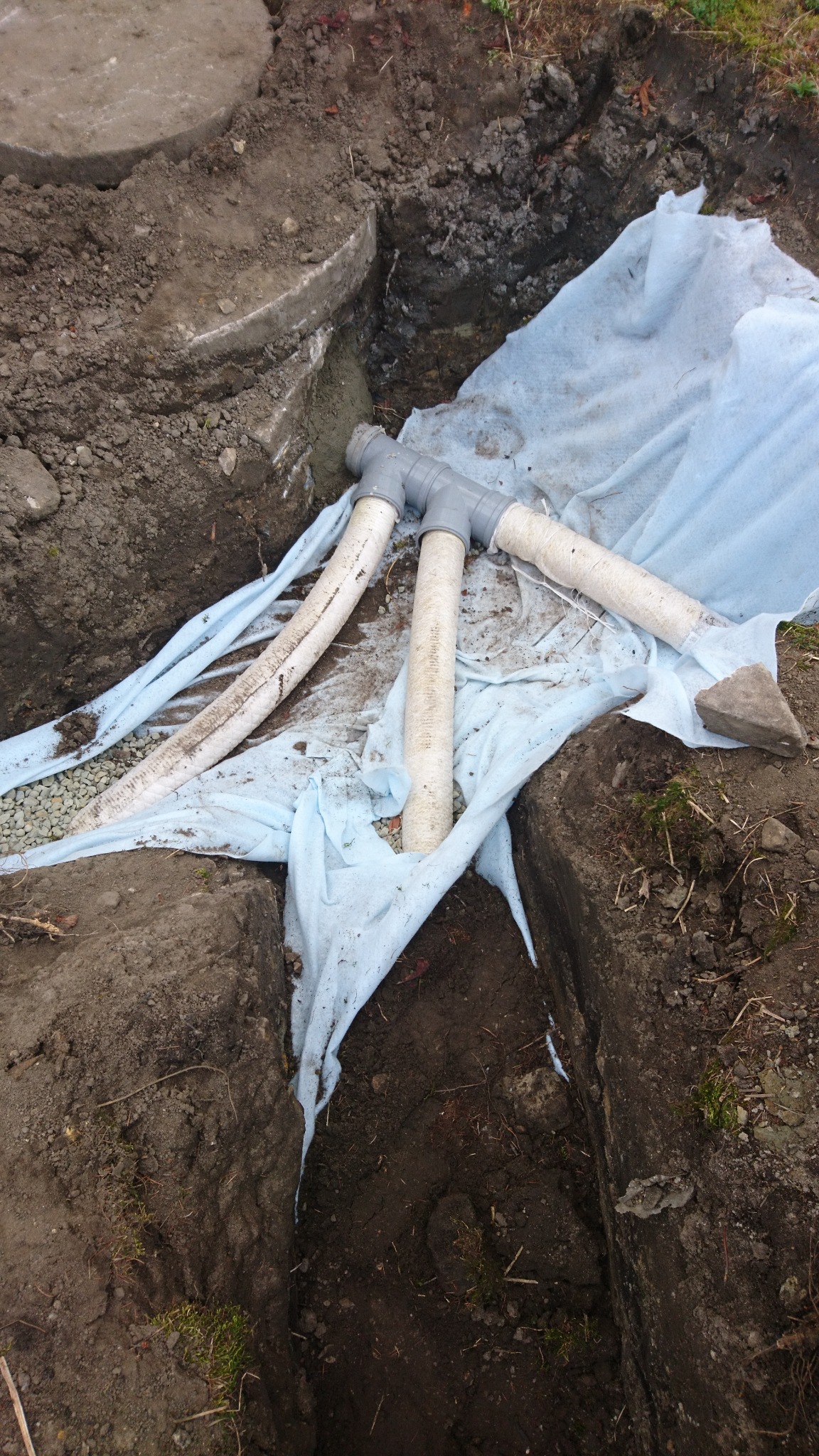 Fragment instalacji przydomowej oczyszczalni ścieków z rurami drenażowymi ułożonymi na geowłókninie w wykopie, widoczne kamienie i ziemia.