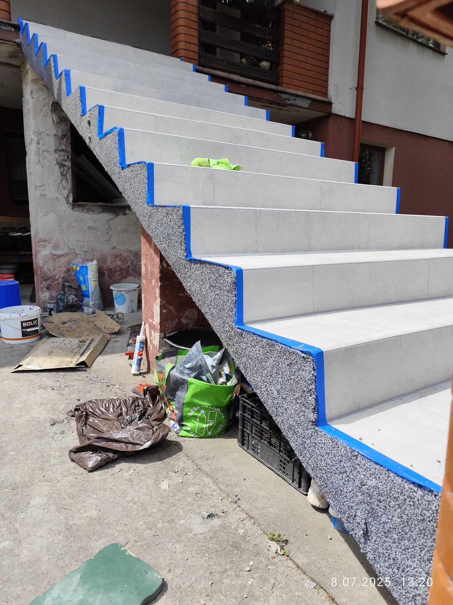 Wykończone schody zewnętrzne z szarym kamieniem dekoracyjnym i jasnymi płytkami, zabezpieczone niebieską taśmą malarską. Widoczne narzędzia i materiały budowlane.