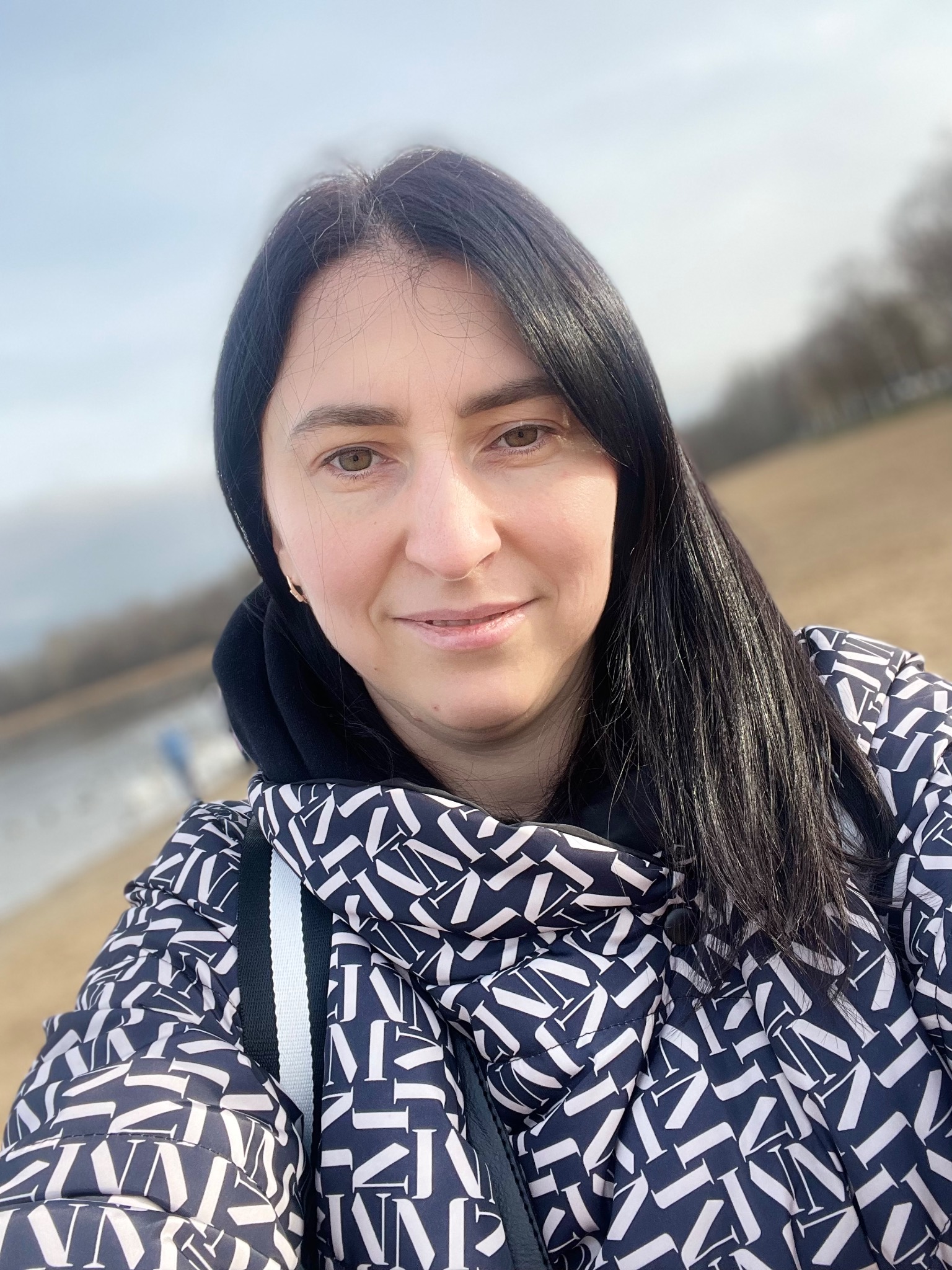 Portret uśmiechniętej kobiety w kurtce z geometrycznym wzorem na tle rozmytego krajobrazu z jeziorem i drzewami.