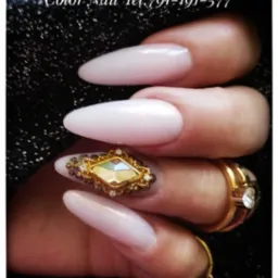 Dłoń z długimi, błyszczącymi paznokciami w odcieniach bieli i różu, jeden z paznokci ozdobiony dużym, złotym ornamentem w kształcie rombu z drobnymi kryształkami, na palcach widoczne złote...