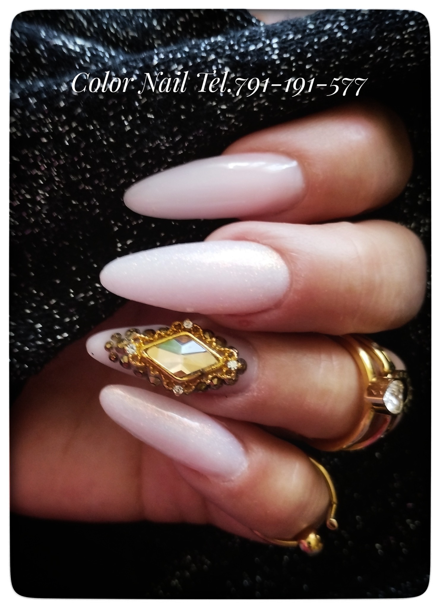 Dłoń z długimi, błyszczącymi paznokciami w odcieniach bieli i różu, jeden z paznokci ozdobiony dużym, złotym ornamentem w kształcie rombu z drobnymi kryształkami, na palcach widoczne złote...