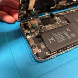 Otwarty iPhone X z widoczną baterią i elementami wewnętrznymi, umieszczony na niebieskiej macie serwisowej z napisem 'Circuit board tool'.