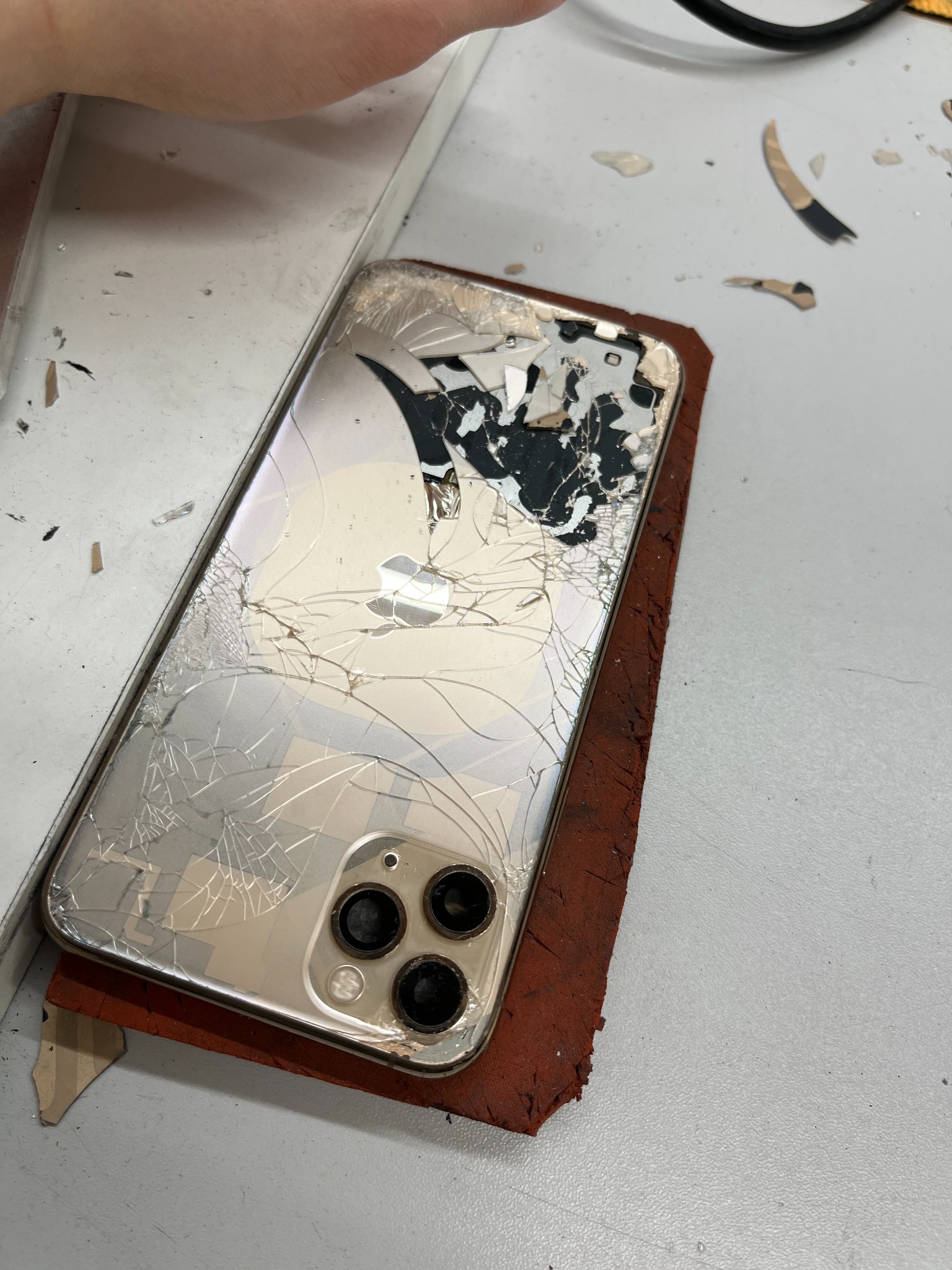 Smartfon z potłuczoną obudową i pękniętym ekranem, leżący na brązowej podkładce, otoczony drobnymi odłamkami szkła.