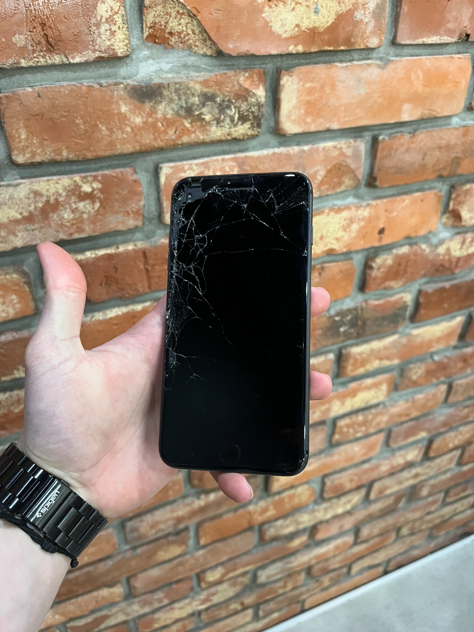 Czarny smartfon z pękniętym ekranem trzymany w dłoni, widoczny metalowy zegarek na nadgarstku, tło z ceglanej ściany.