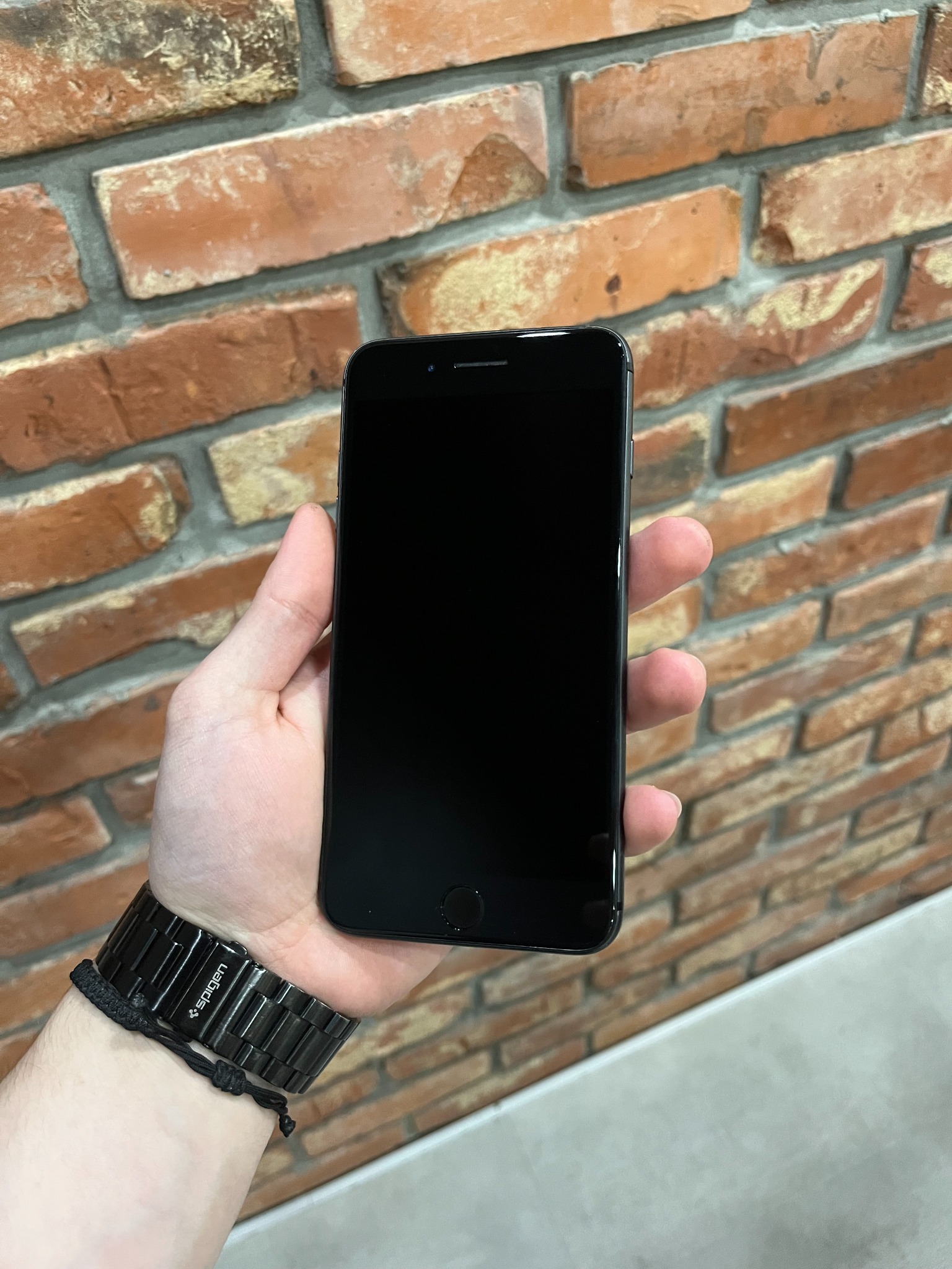 Czarny smartfon trzymany w dłoni, na tle ściany z cegły. Widoczny metalowy pasek do zegarka marki Spigen.