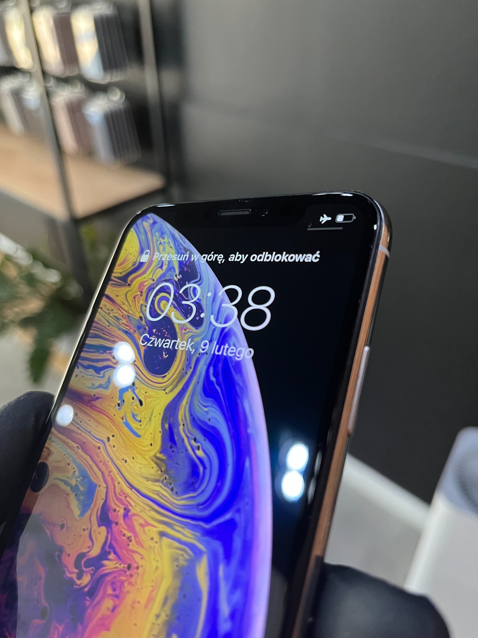 Smartfon iPhone z włączonym ekranem blokady, wyświetlający godzinę 03:38 w czwartek, 9 lutego, z aktywnym trybem samolotowym i niskim poziomem baterii, trzymany w rękawicy ochronnej.