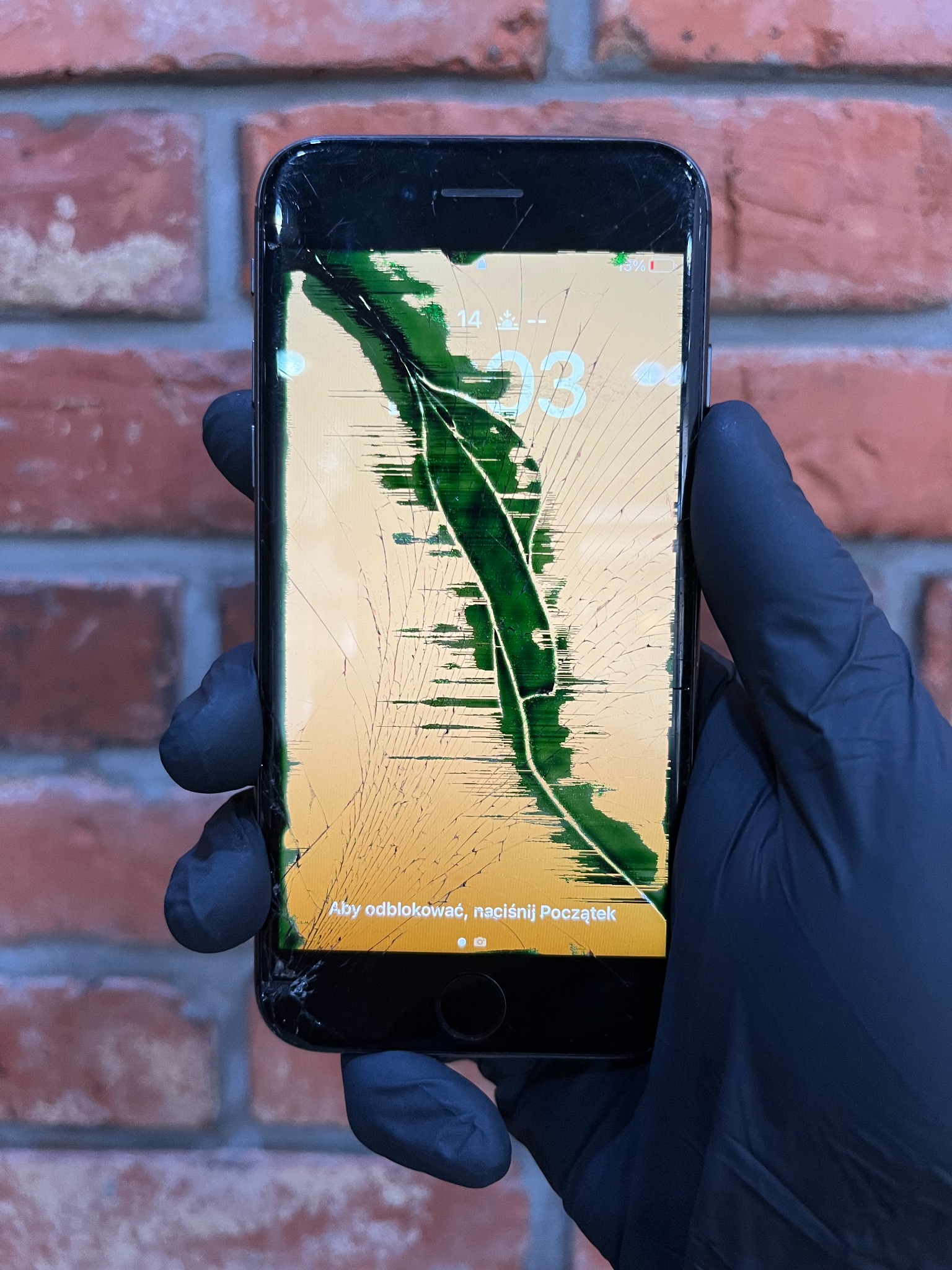 Smartfon z całkowicie pękniętym ekranem, trzymany w dłoni w czarnej rękawiczce na tle ceglanej ściany. Uszkodzenie wygląda jak rozgałęziające się korzenie drzewa.