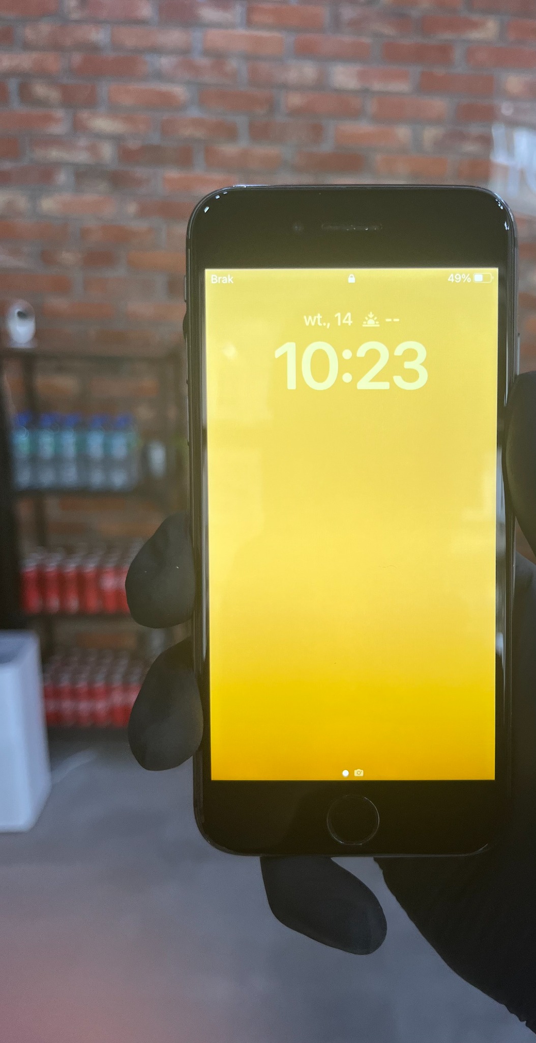 Czarny smartfon z żółtym ekranem wyświetlającym godzinę 10:23, trzymany w dłoni w czarnej rękawiczce na tle ceglanej ściany i częściowo widocznych półek z napojami.