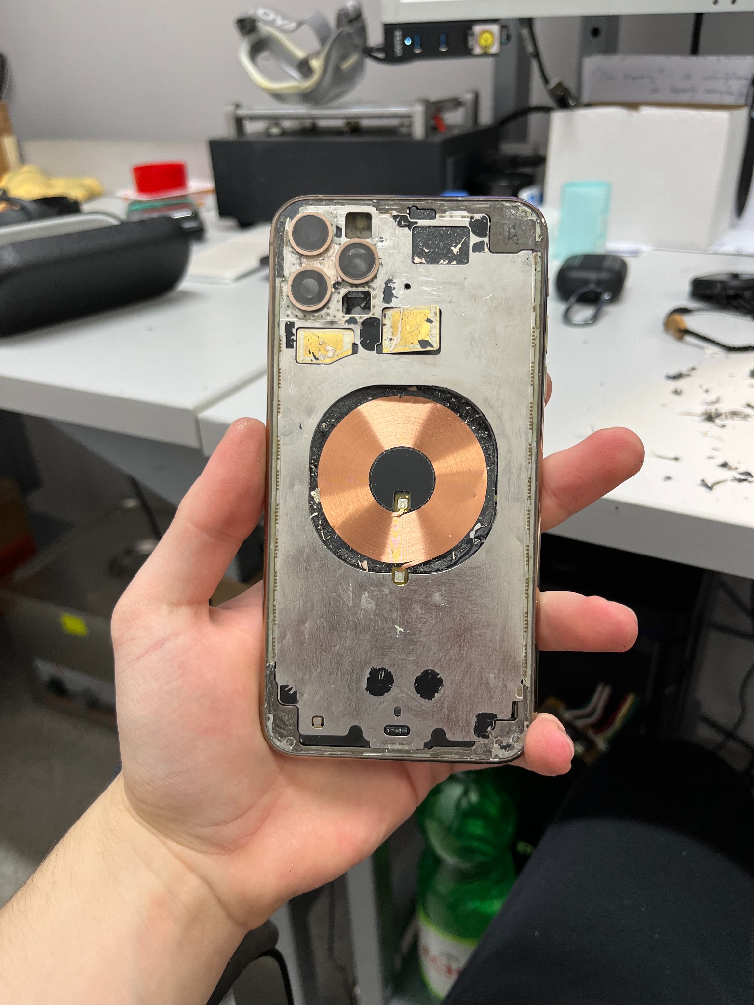 Rozłożony iPhone 13 Pro Max w dłoni, widoczna tylna obudowa bez szkła, z miedzianą cewką ładowania bezprzewodowego i otworami na kamery, na tle warsztatu z narzędziami.