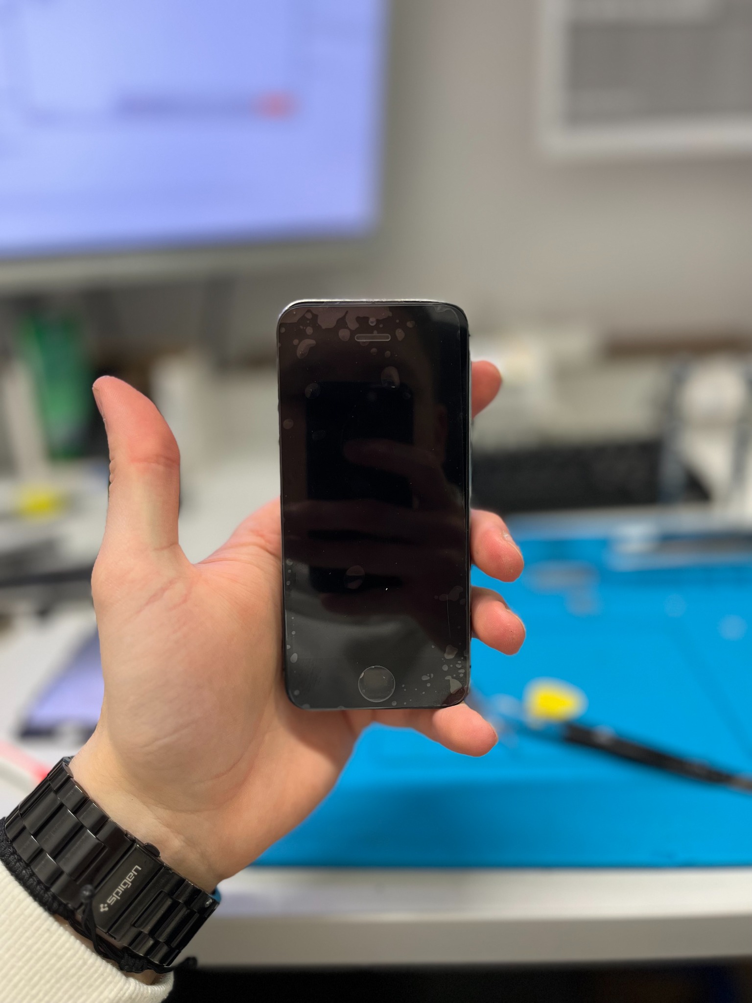 Czarny smartfon z uszkodzonym ekranem w dłoni, widoczne ślady po kleju i pęknięcia, na tle mata serwisowa i narzędzia.