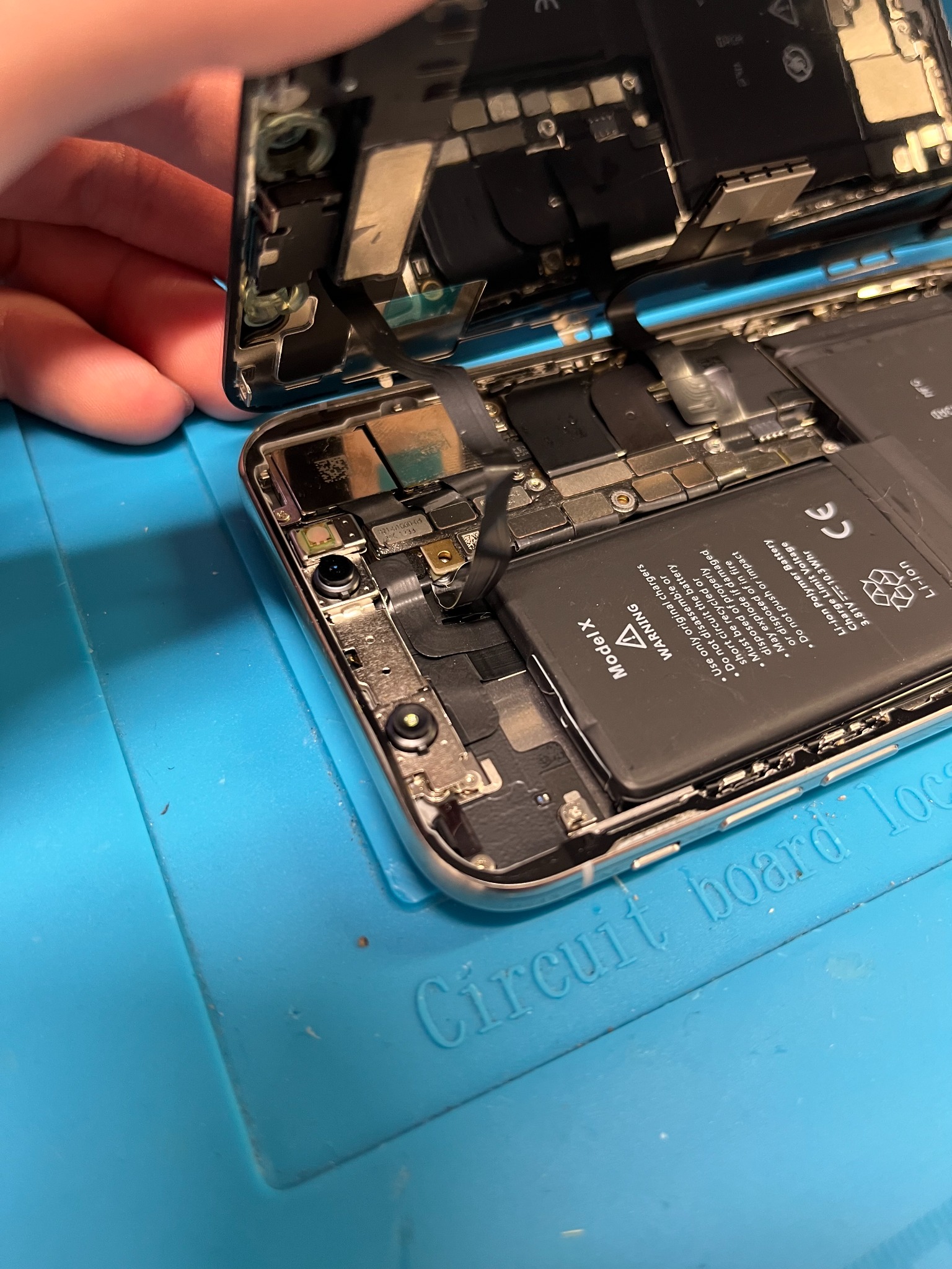 Otwarty iPhone X z widoczną baterią i elementami wewnętrznymi, umieszczony na niebieskiej macie serwisowej z napisem 'Circuit board tool'.