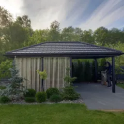 Nowoczesna konstrukcja z drewnianymi lamelami i grafitowym dachem, prawdopodobnie pergola, otoczona zielenią i kostką brukową, z pracownikiem w tle wykonującym prace wykończeniowe.