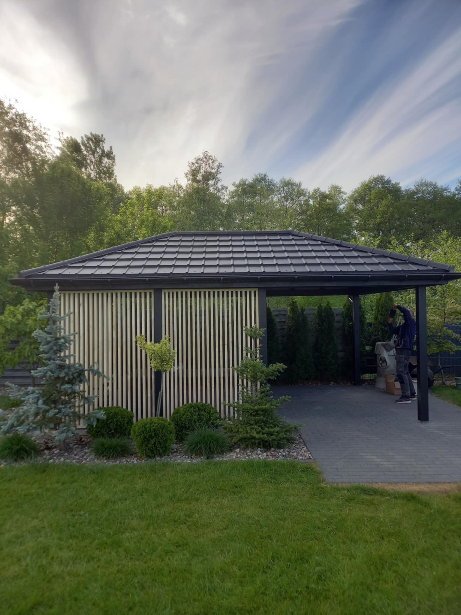 Nowoczesna konstrukcja z drewnianymi lamelami i grafitowym dachem, prawdopodobnie pergola, otoczona zielenią i kostką brukową, z pracownikiem w tle wykonującym prace wykończeniowe.