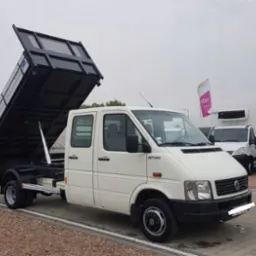 Biały samochód ciężarowy z otwartą, czarną skrzynią ładunkową na podwoziu, widoczny na parkingu z kostki brukowej.