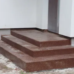 Brązowe, trzystopniowe schody wejściowe z wykończeniem kamyczkowym, prowadzące do budynku z białymi ścianami, widoczne z bliskiej perspektywy.