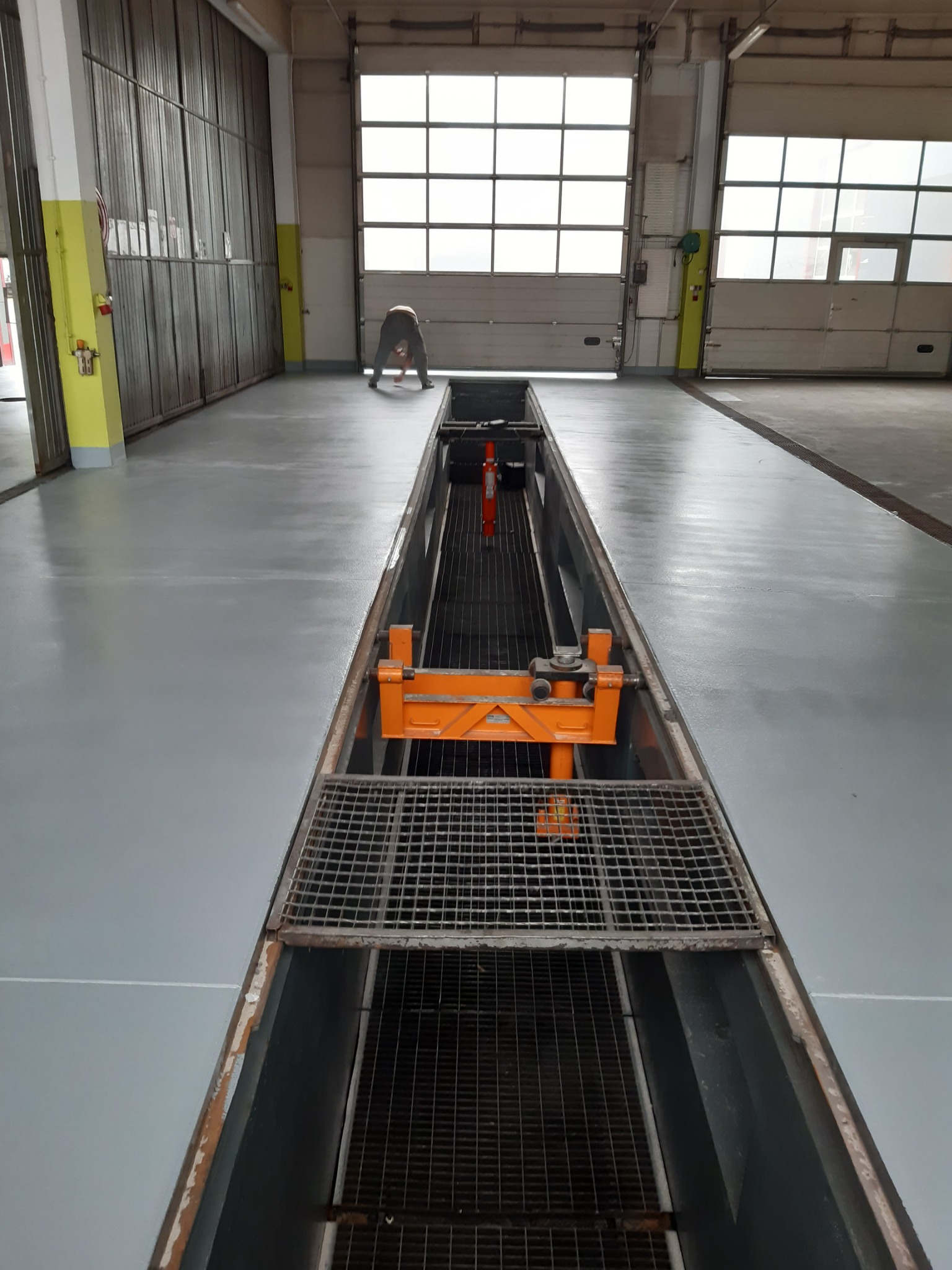 Szara, gładka podłoga w warsztacie samochodowym z kanałem naprawczym i pomarańczowym podnośnikiem, widoczna osoba w tle.