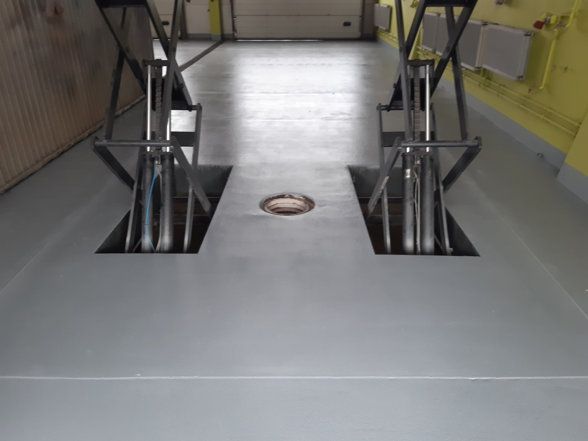 Szara, gładka posadzka żywiczna w warsztacie samochodowym z wbudowanymi podnośnikami, widoczne otwory i metalowe konstrukcje podnośników.