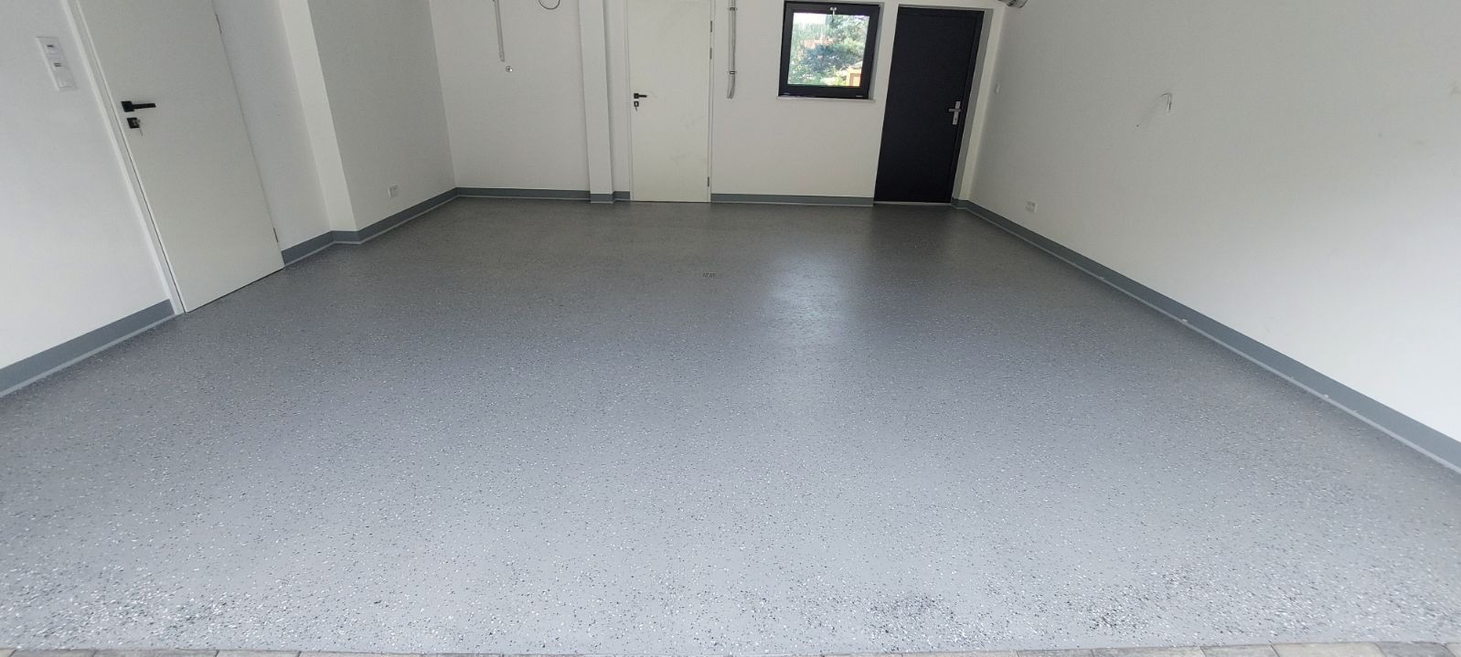 Szara, gładka podłoga z drobnymi, ciemniejszymi wtrąceniami w jasnym pomieszczeniu z trzema parami drzwi i małym oknem.