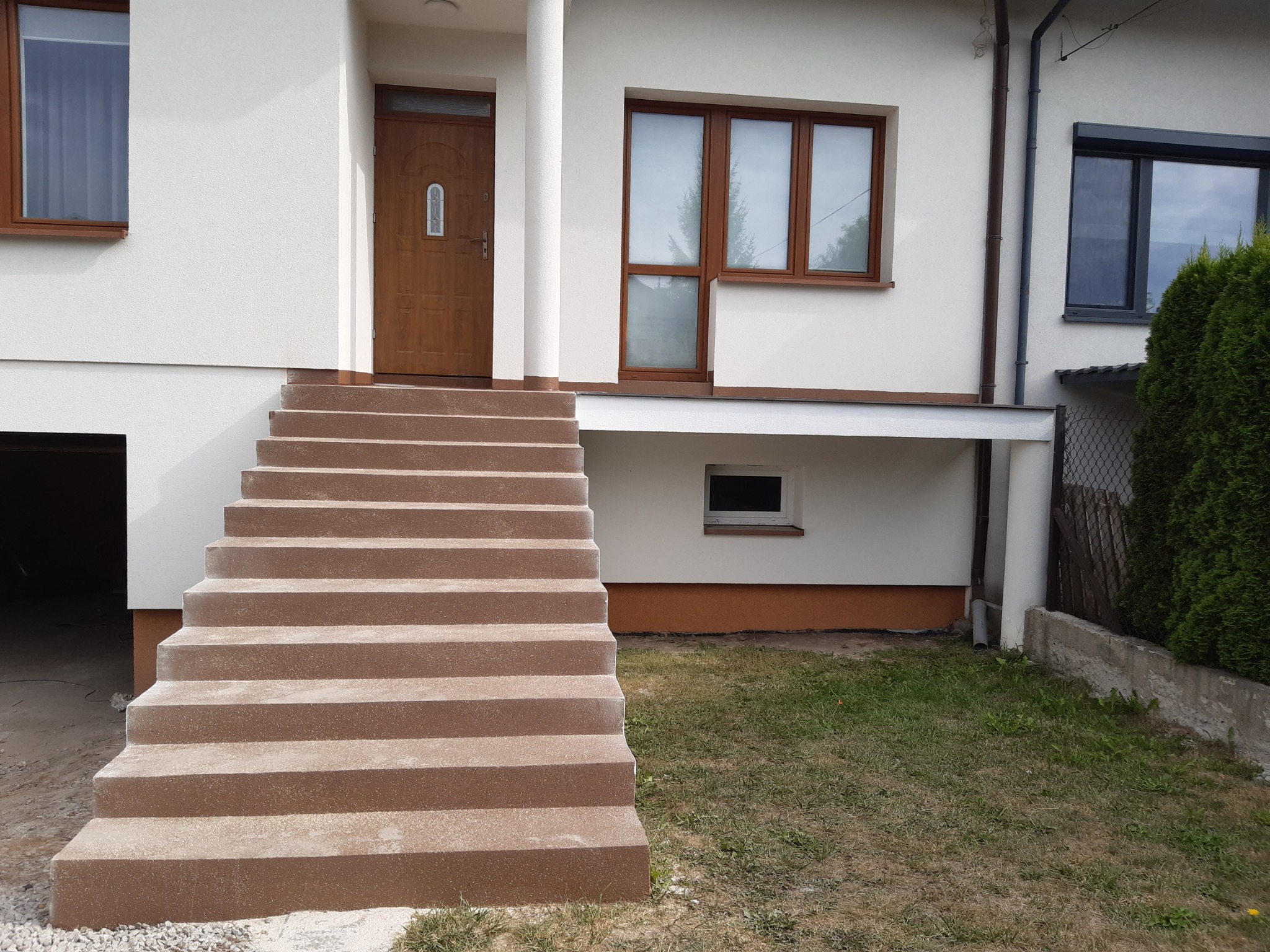 Zewnętrzne schody wejściowe do domu, pokryte brązową, chropowatą powłoką, widok z dołu na tle białej elewacji budynku z brązowymi oknami i drzwiami.