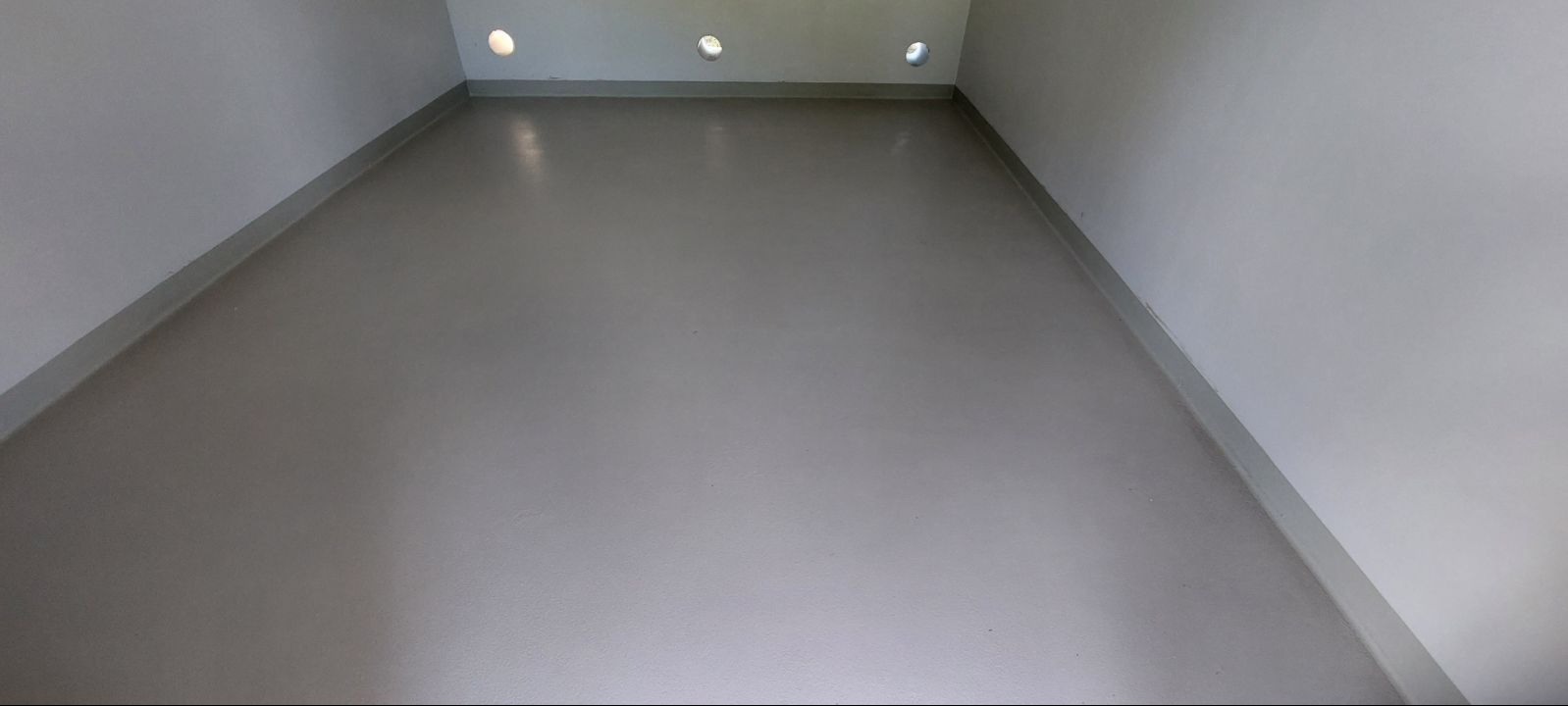 Gładka, szara posadzka żywiczna w pomieszczeniu z białymi ścianami i listwami przypodłogowymi, oświetlona przez okrągłe otwory w górnej części ściany.