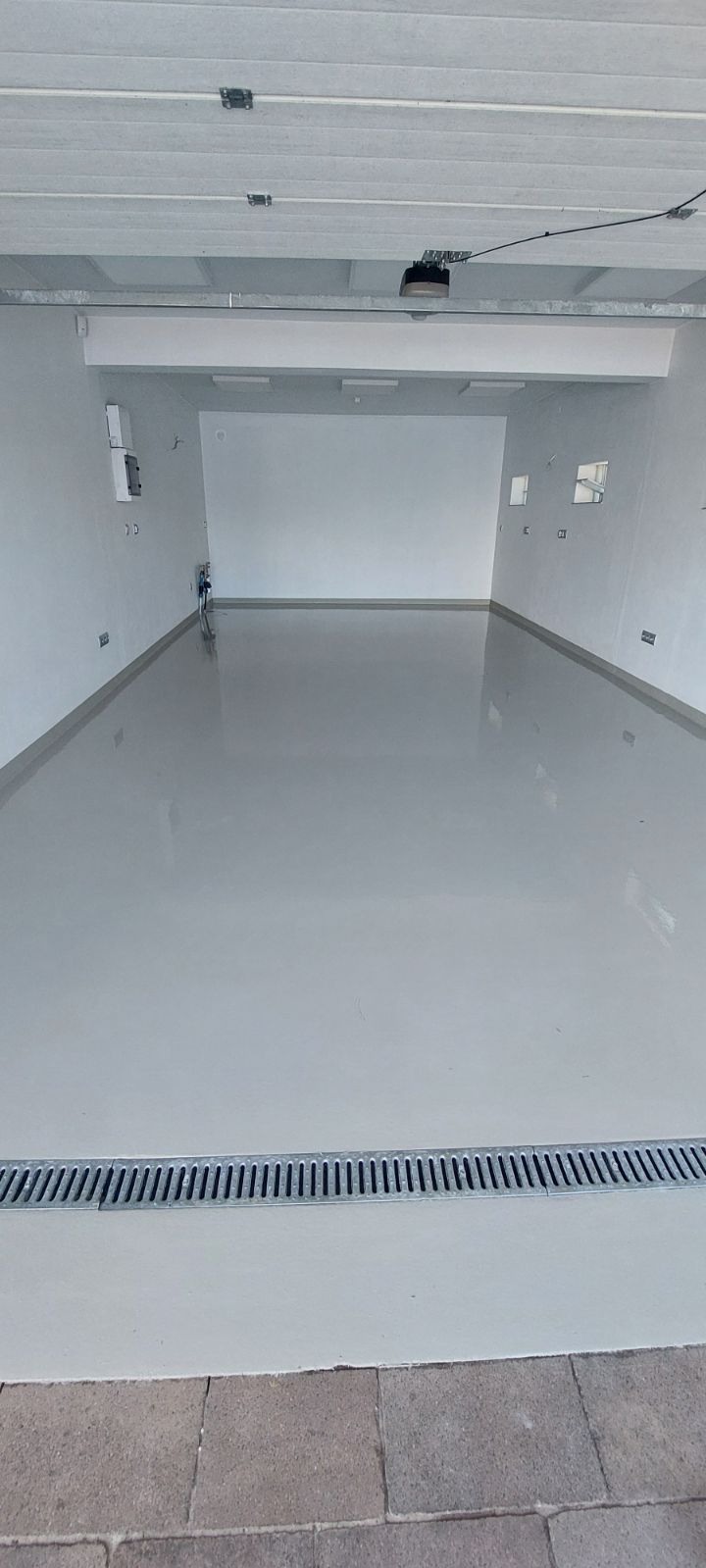 Jasnoszara, gładka podłoga w garażu z odpływem liniowym przy bramie, widok z zewnątrz.