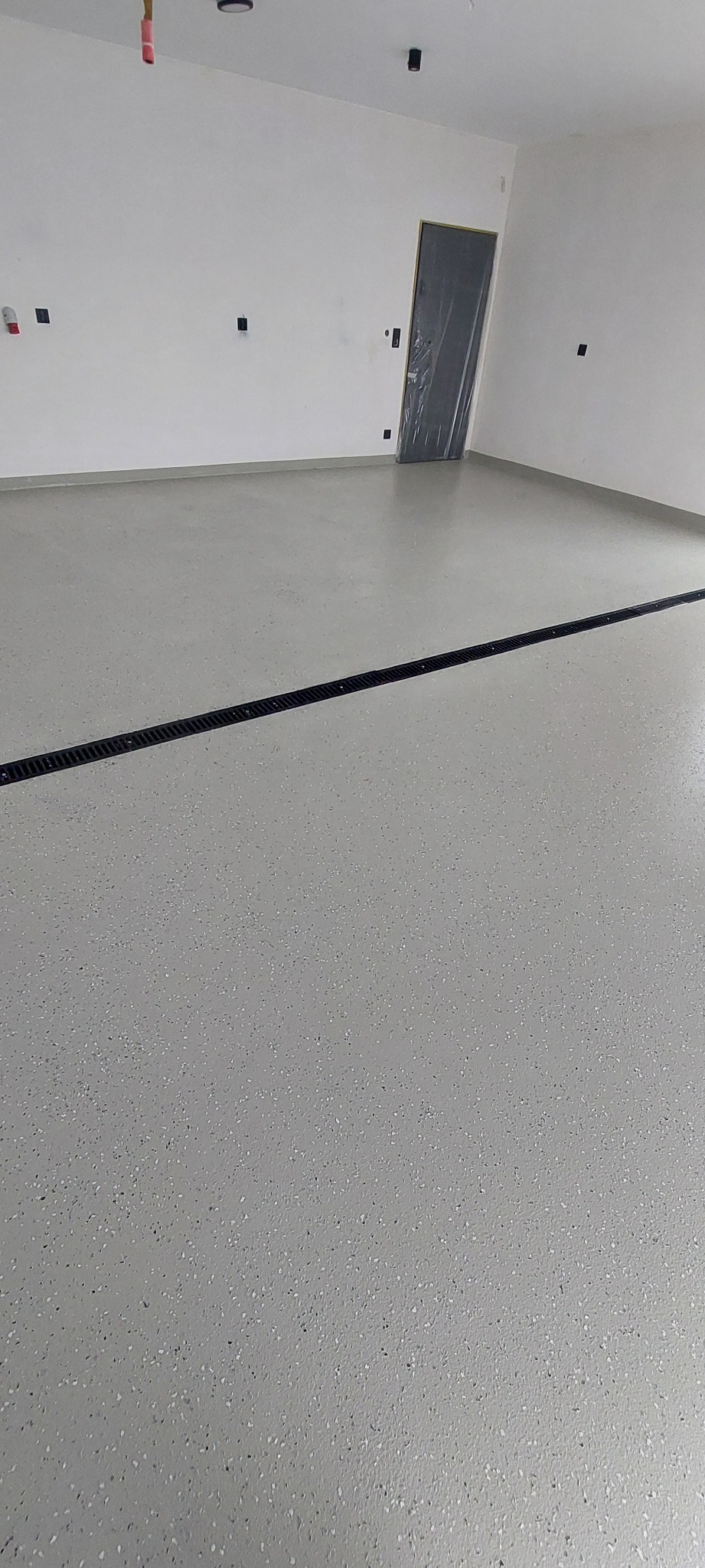 Jasnoszara, błyszcząca posadzka z drobnymi, ciemnymi inkluzjami w nowym pomieszczeniu, z widocznym liniowym odpływem podłogowym.