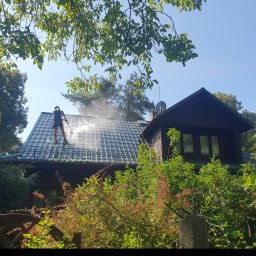 MW Team - Pracownik w kombinezonie czyści dach z szarej dachówki za pomocą myjki ciśnieniowej, w tle dom z ciemnego drewna i bujna roślinność.
