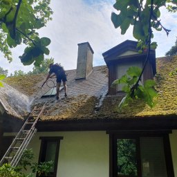 MW Team - Pracownik w uprzęży, z drabiny spryskuje dach z mchu wodą pod ciśnieniem w miejscowości Stare Babice. Widoczny komin i okno dachowe.