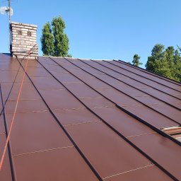 MW Team - Ciemnobrązowy dach z blachy na domu jednorodzinnym, widoczny komin z anteną i liną zabezpieczającą, drzewa w tle, błękitne niebo.
