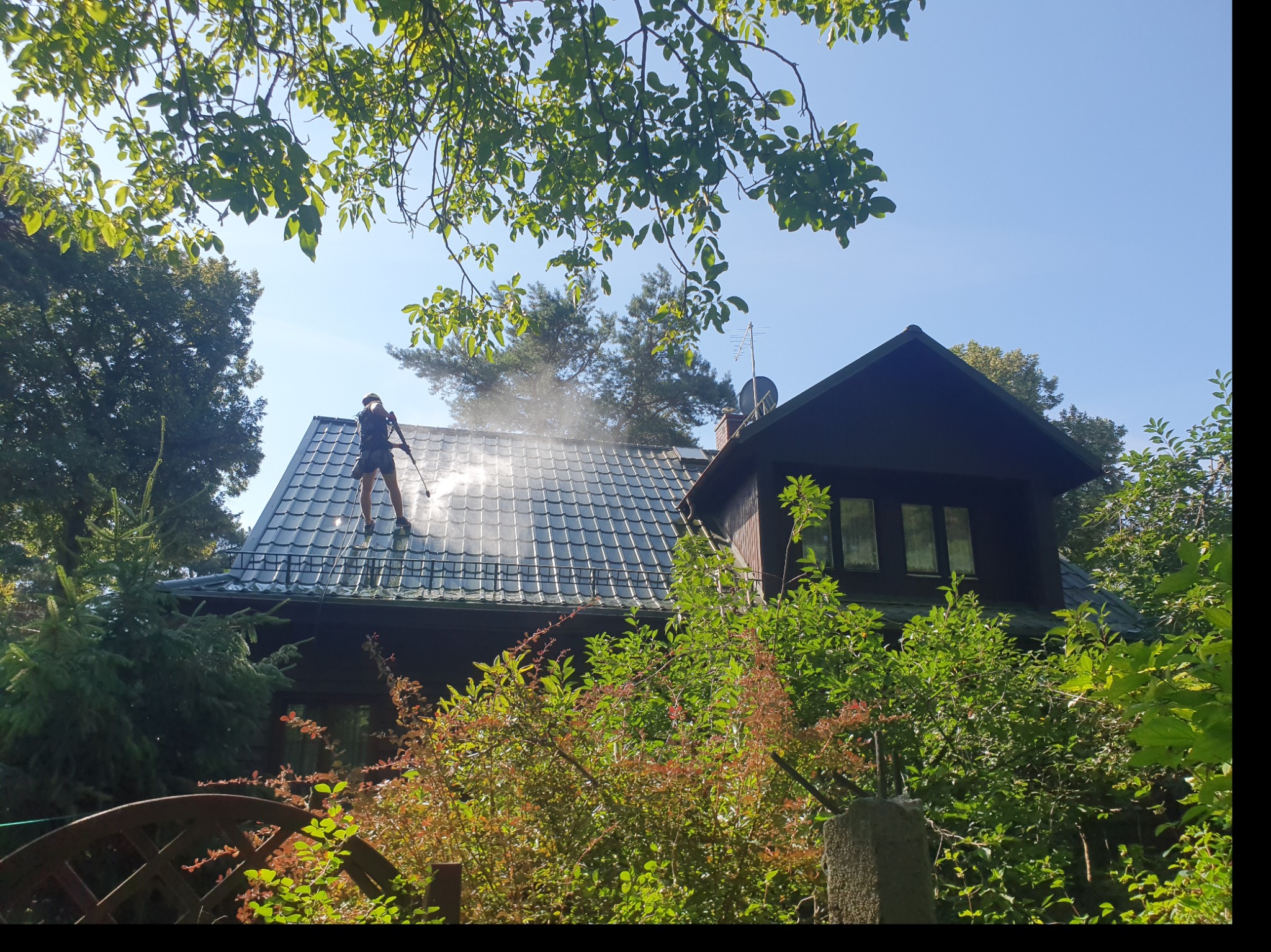 Pracownik w kombinezonie czyści dach z szarej dachówki za pomocą myjki ciśnieniowej, w tle dom z ciemnego drewna i bujna roślinność.