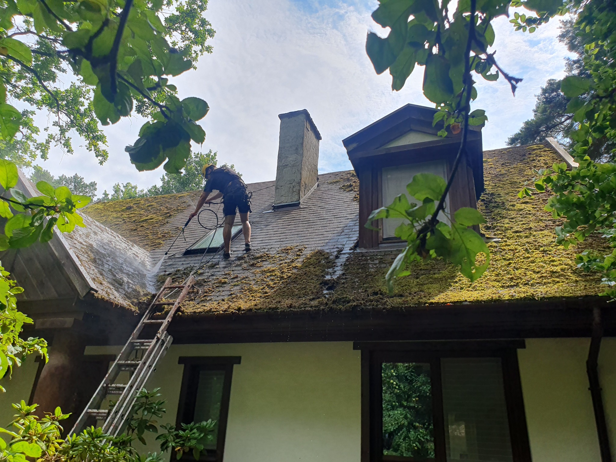 Pracownik w uprzęży, z drabiny spryskuje dach z mchu wodą pod ciśnieniem w miejscowości Stare Babice. Widoczny komin i okno dachowe.