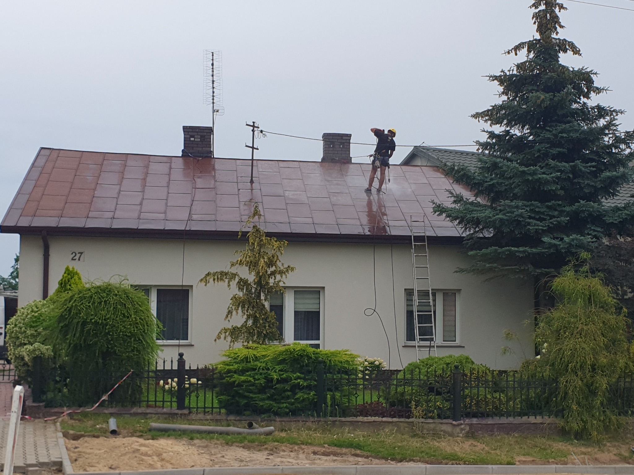 Pracownik w uprzęży, zraszaczem ciśnieniowym, myje dach domu jednorodzinnego z brązowej dachówki, obok komin, antena i drabina oparta o ścianę.