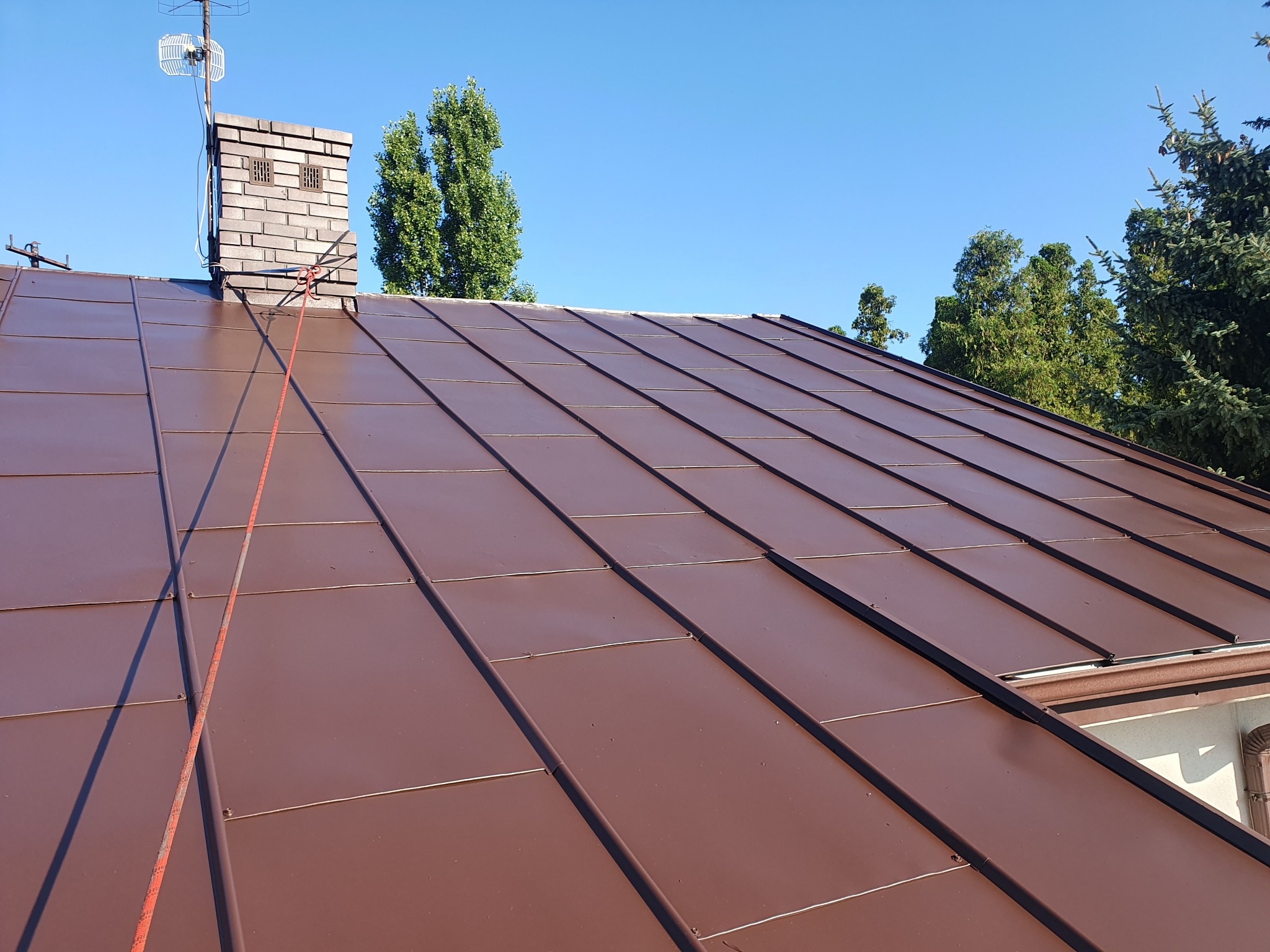 Ciemnobrązowy dach z blachy na domu jednorodzinnym, widoczny komin z anteną i liną zabezpieczającą, drzewa w tle, błękitne niebo.